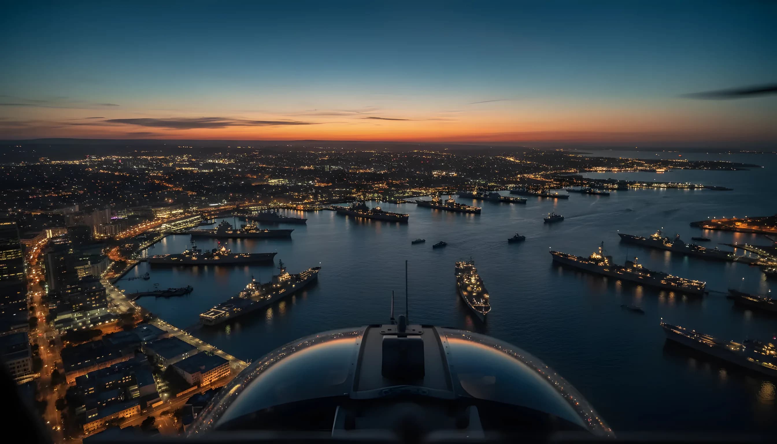 Aerial view of a naval fleet in a city harbor at dusk, seen from an approaching helicopter.