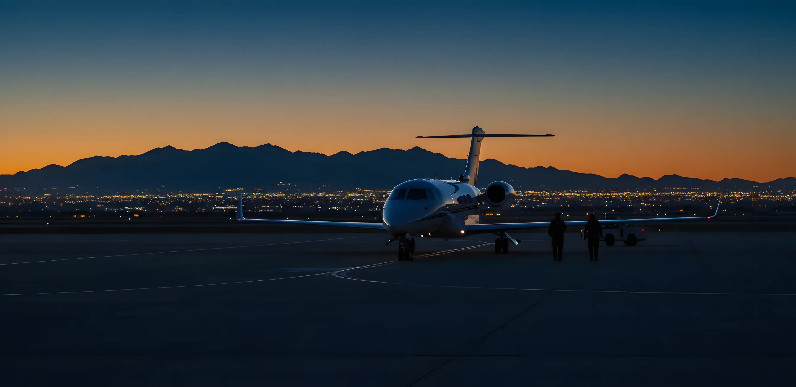 A private jet on an airport tarmac at twilight, with mountains and distant city lights in the background.