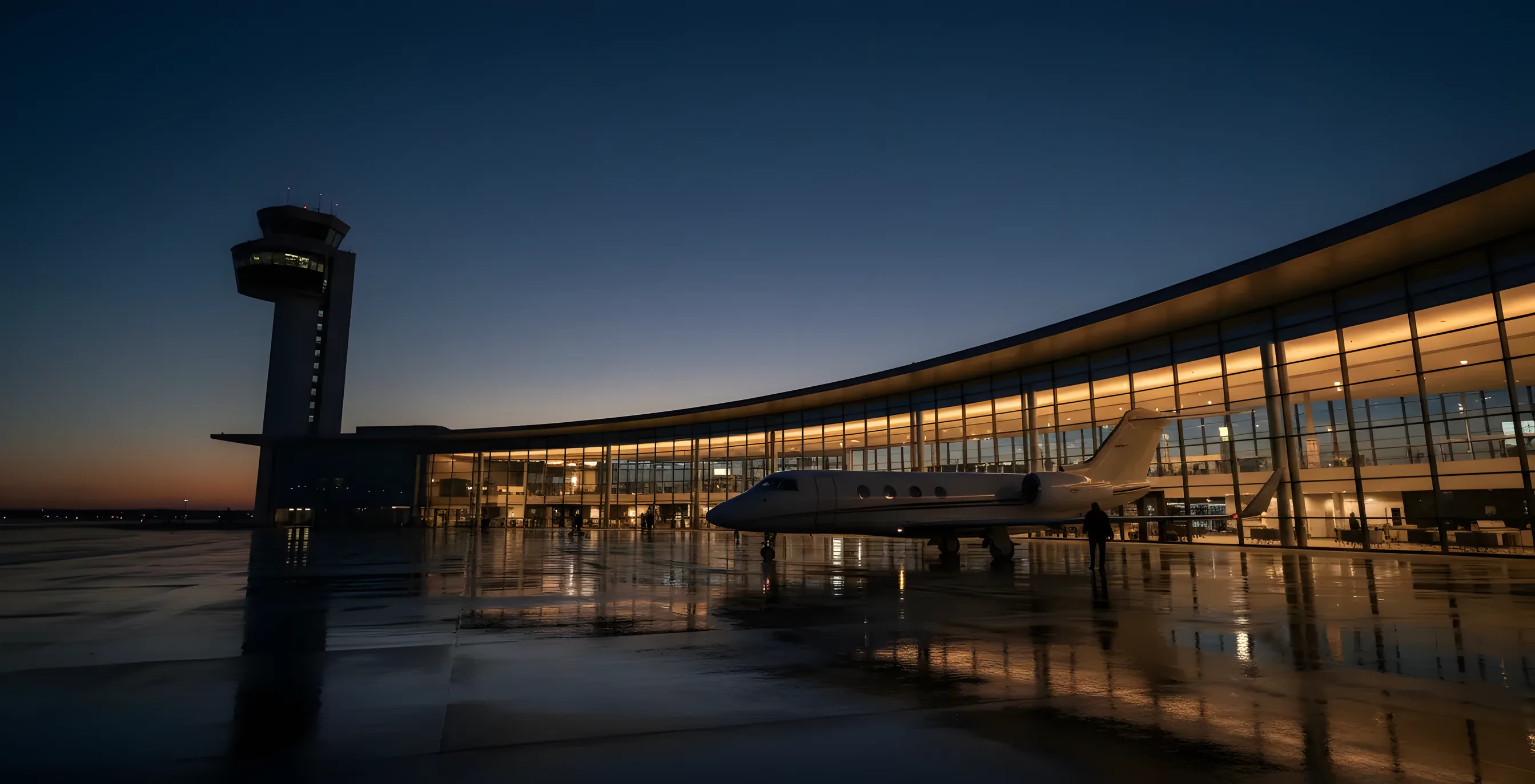 A modern airport terminal and control tower at dusk, with warm lights glowing from within.