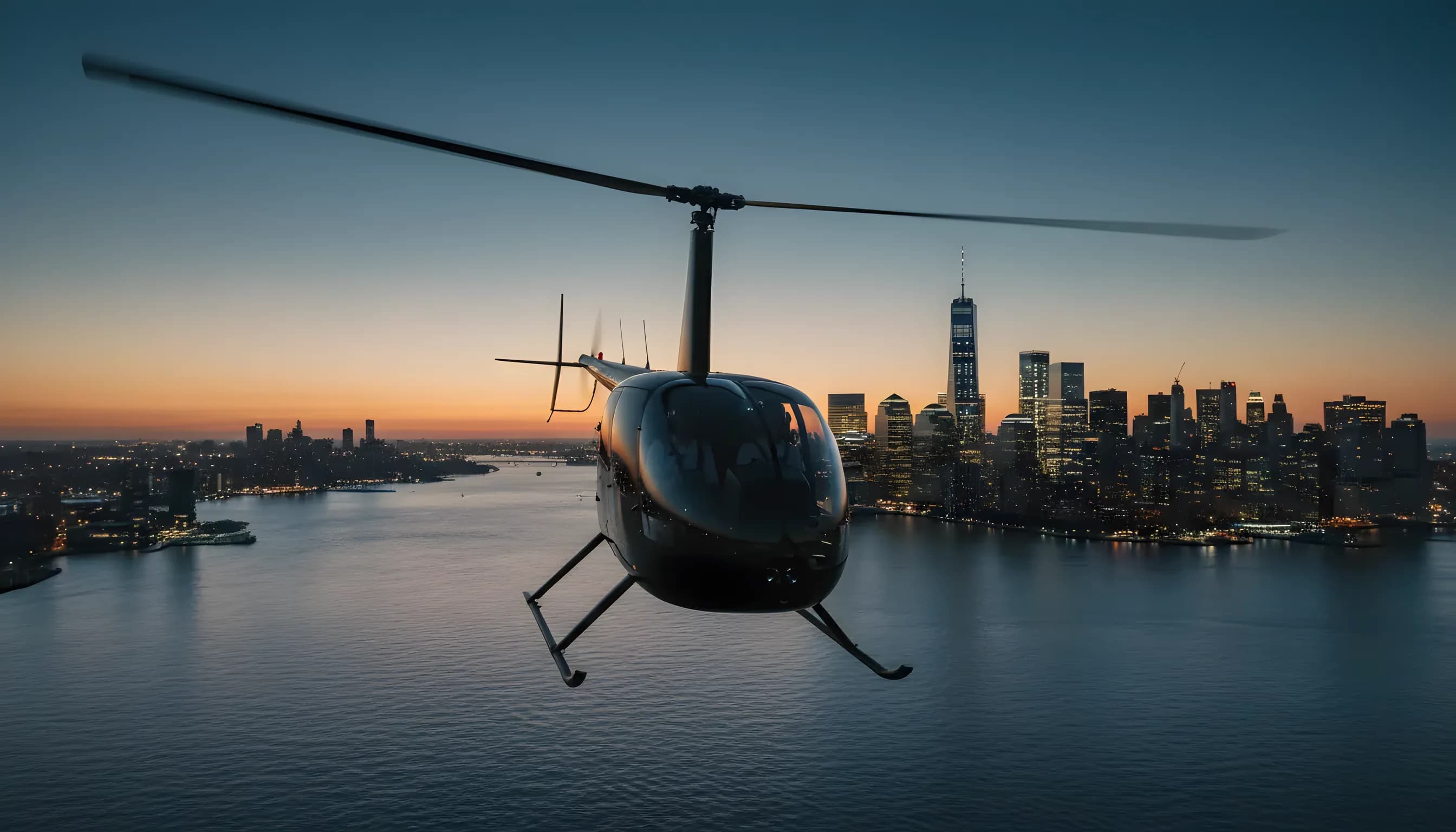 A helicopter flies over the water towards the illuminated Lower Manhattan skyline just after sunset.