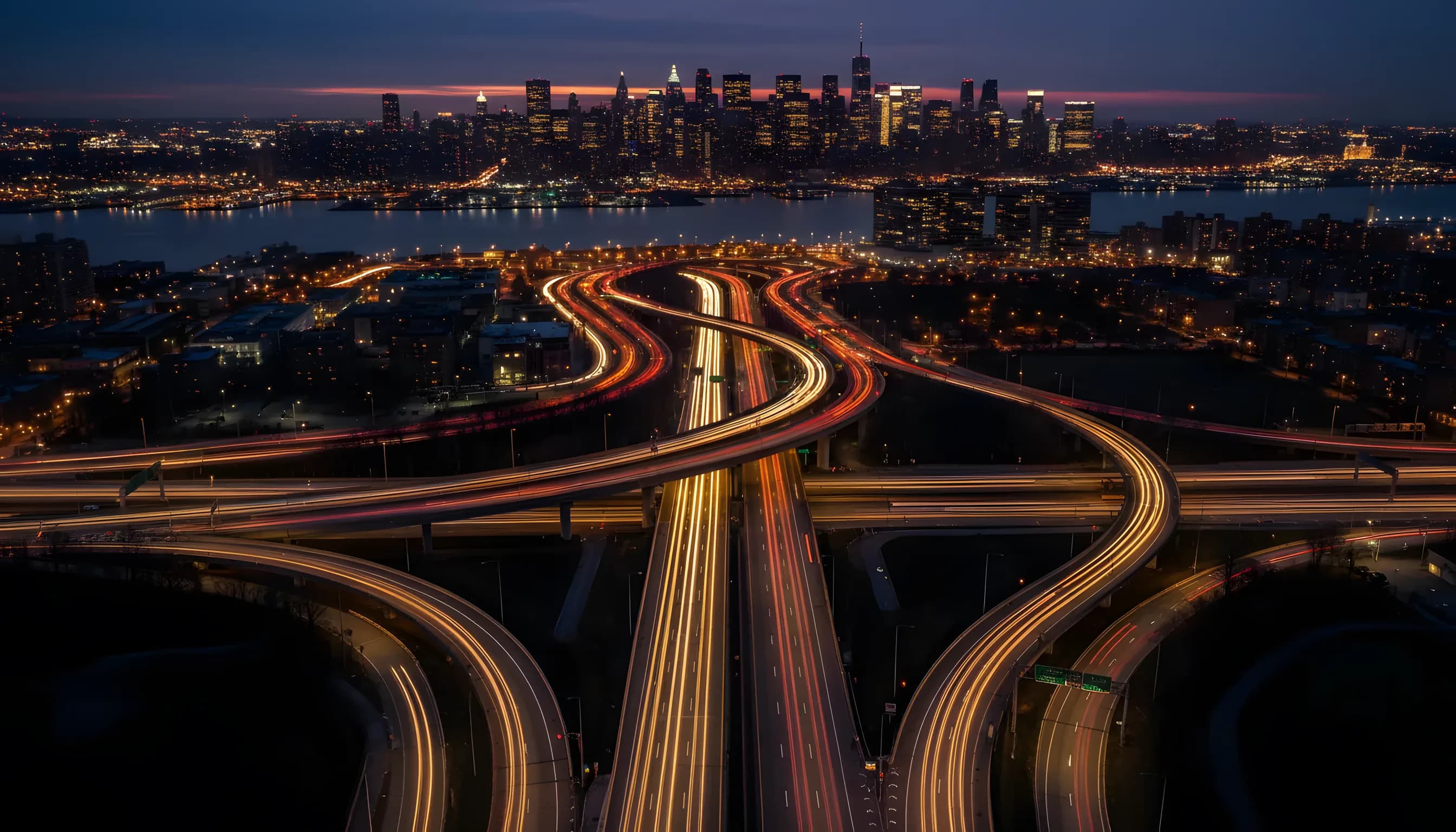 Aerial view of the highway network in New Jersey at dusk, with the illuminated Manhattan skyline in the distance.