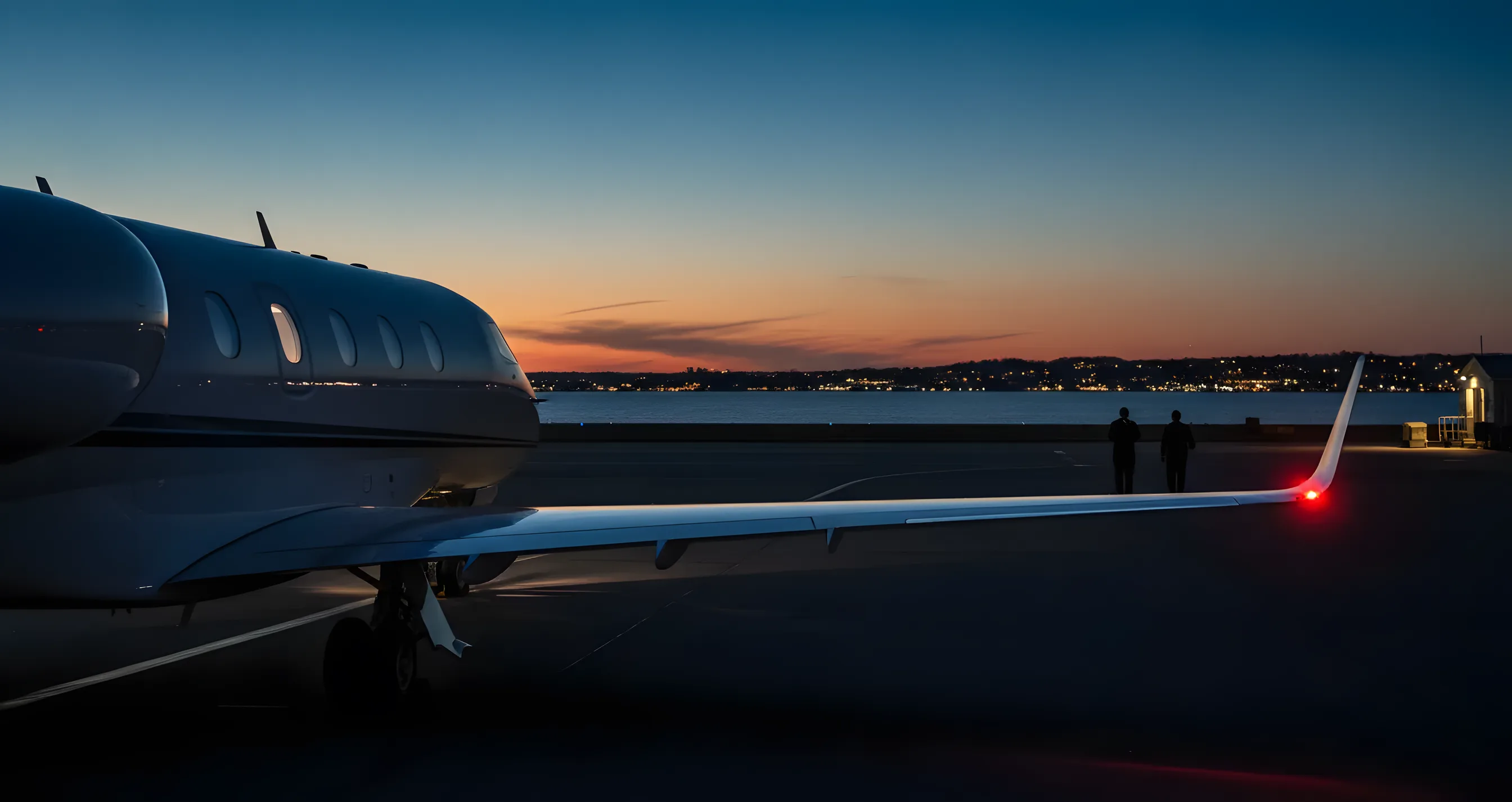 A private jet on an airport tarmac at dusk, with the lights of a coastal city visible across the water.