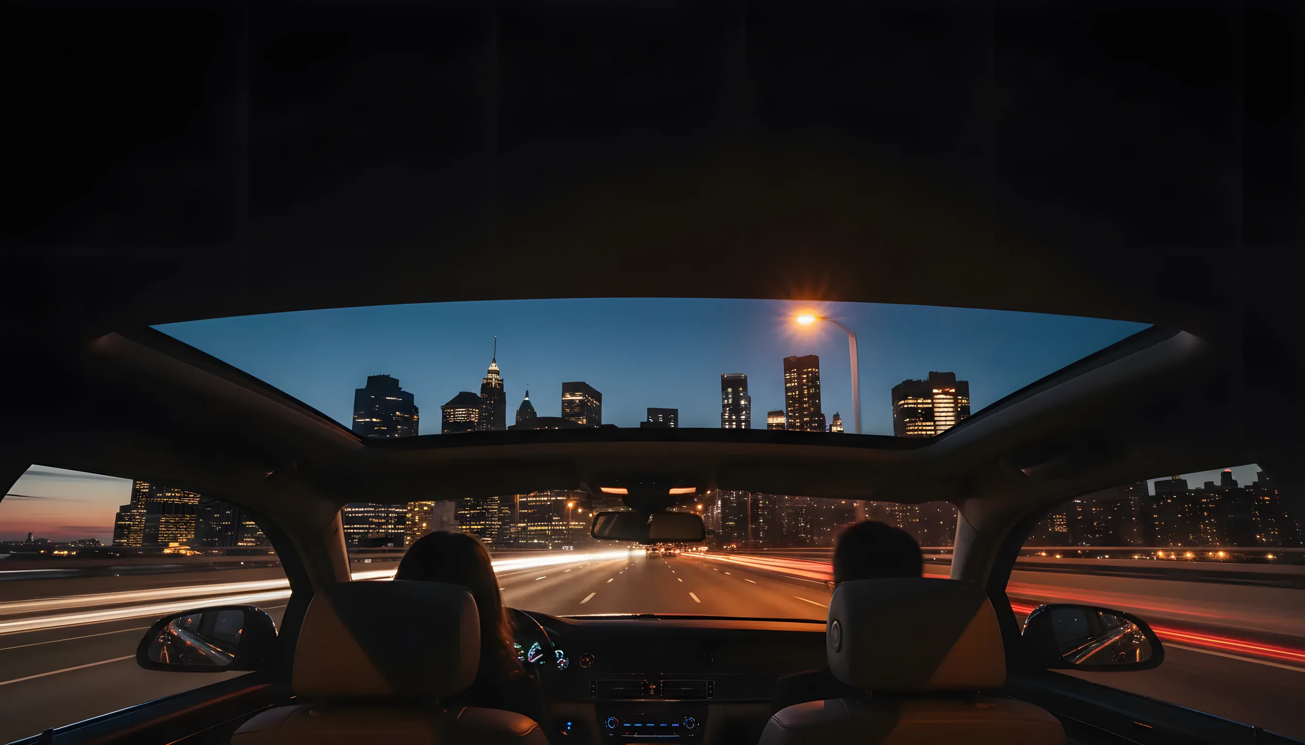 The Manhattan skyline glows with amber lights at dusk, viewed from inside a car driving over a bridge.