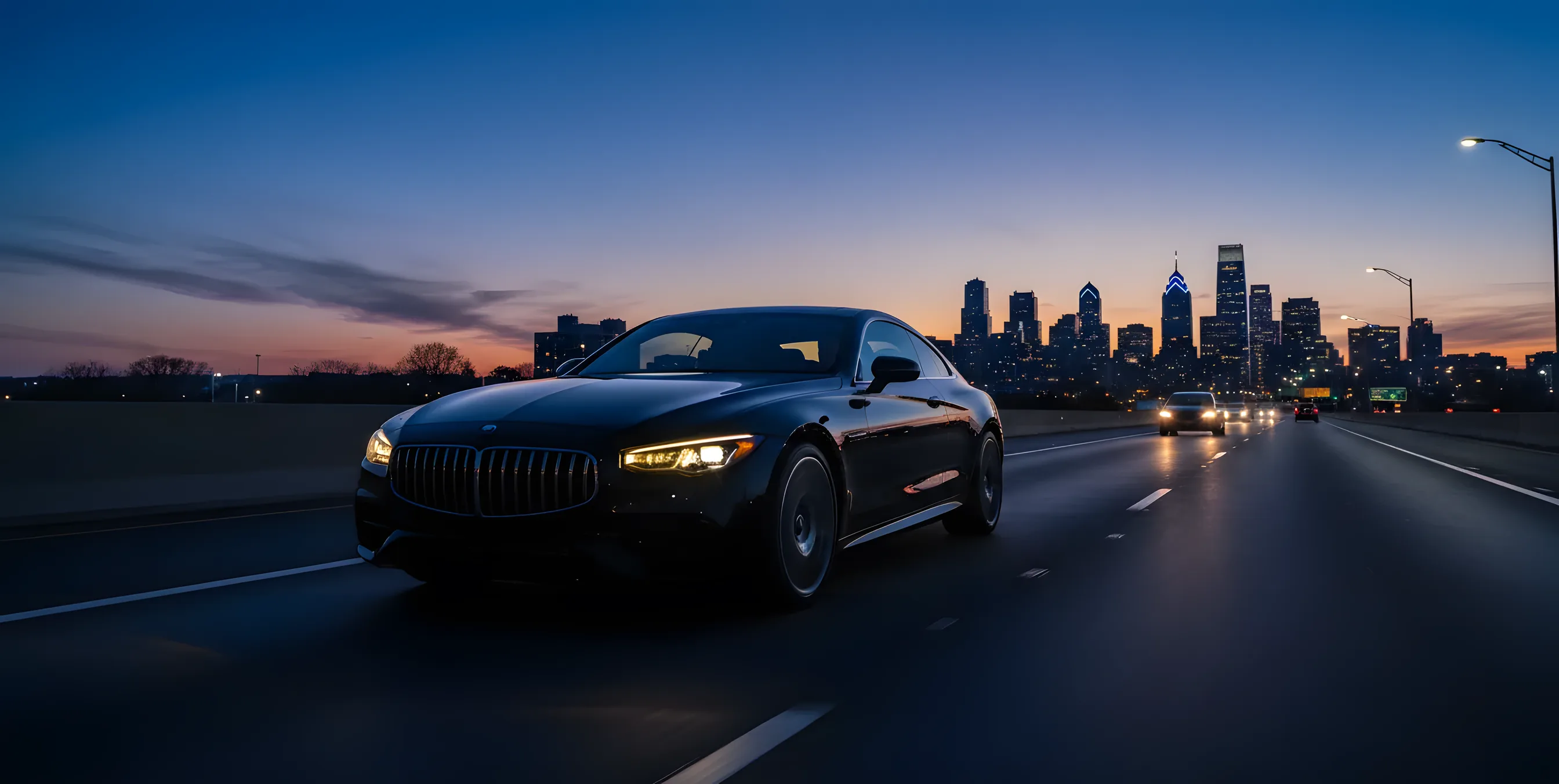 A luxury sedan drives on a highway at dusk, with a city skyline glowing in the distance.