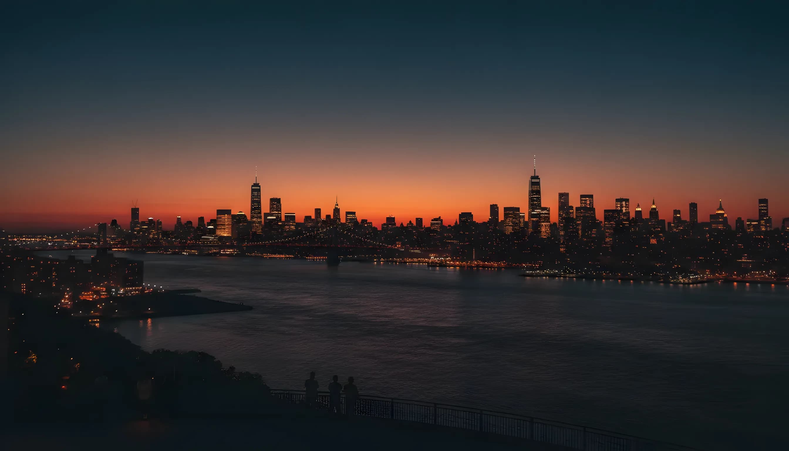 The Manhattan skyline glows with amber lights at dusk, viewed from across the river in New Jersey.