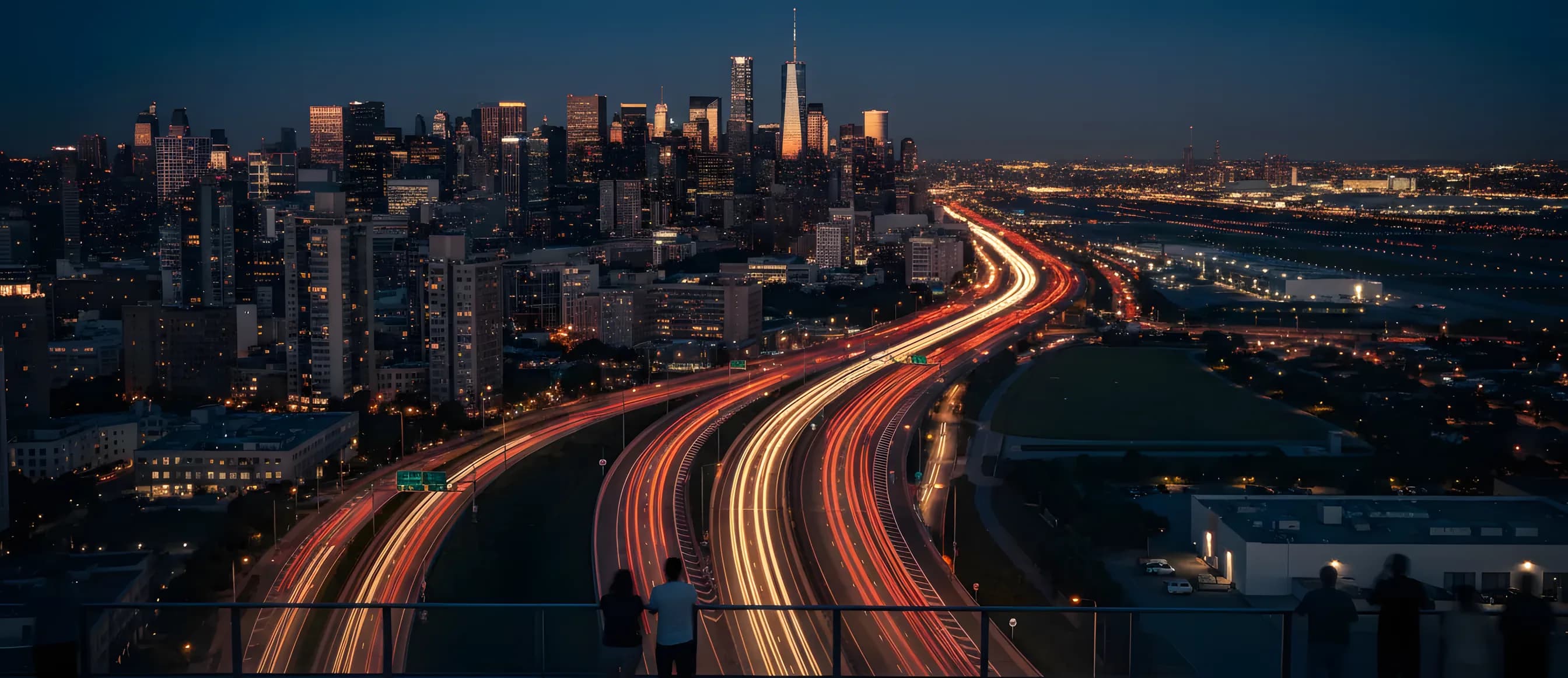 A city skyline at dusk with light trails from cars on a highway leading towards a distant airport.