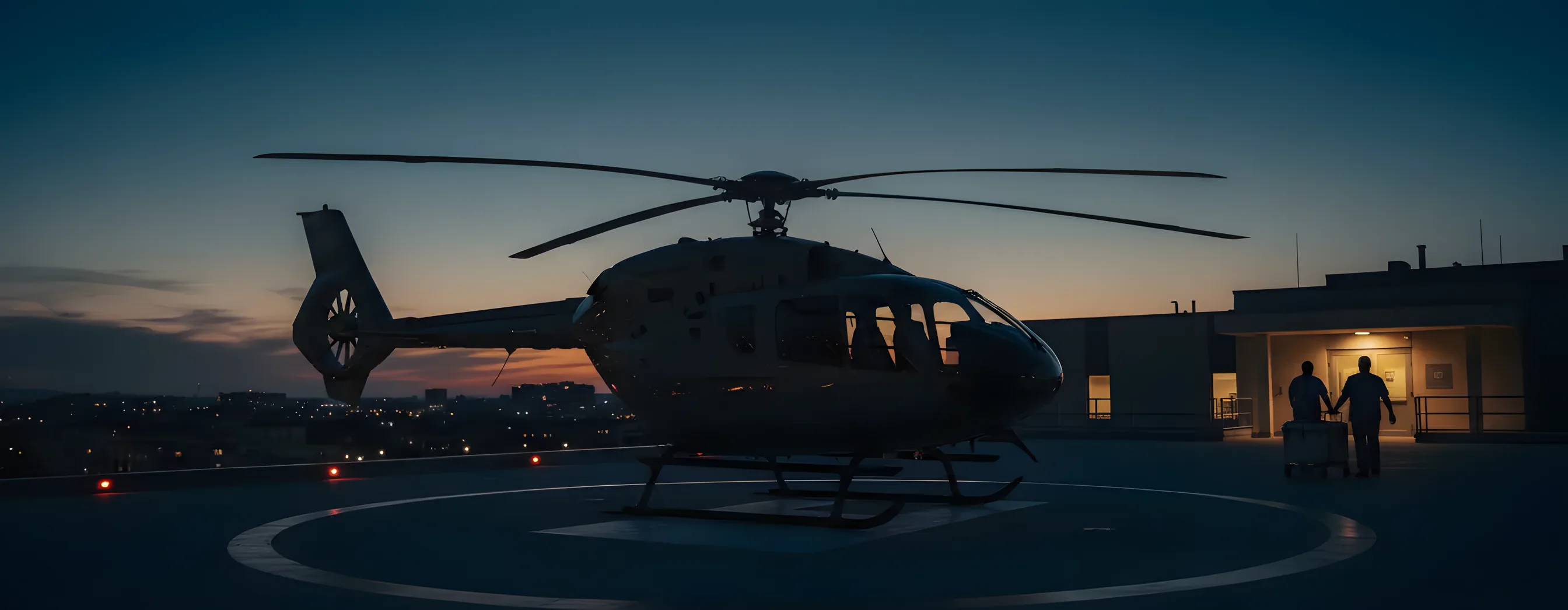 A helicopter on a hospital rooftop helipad at dusk as two staff wheel a transport cooler from the building.
