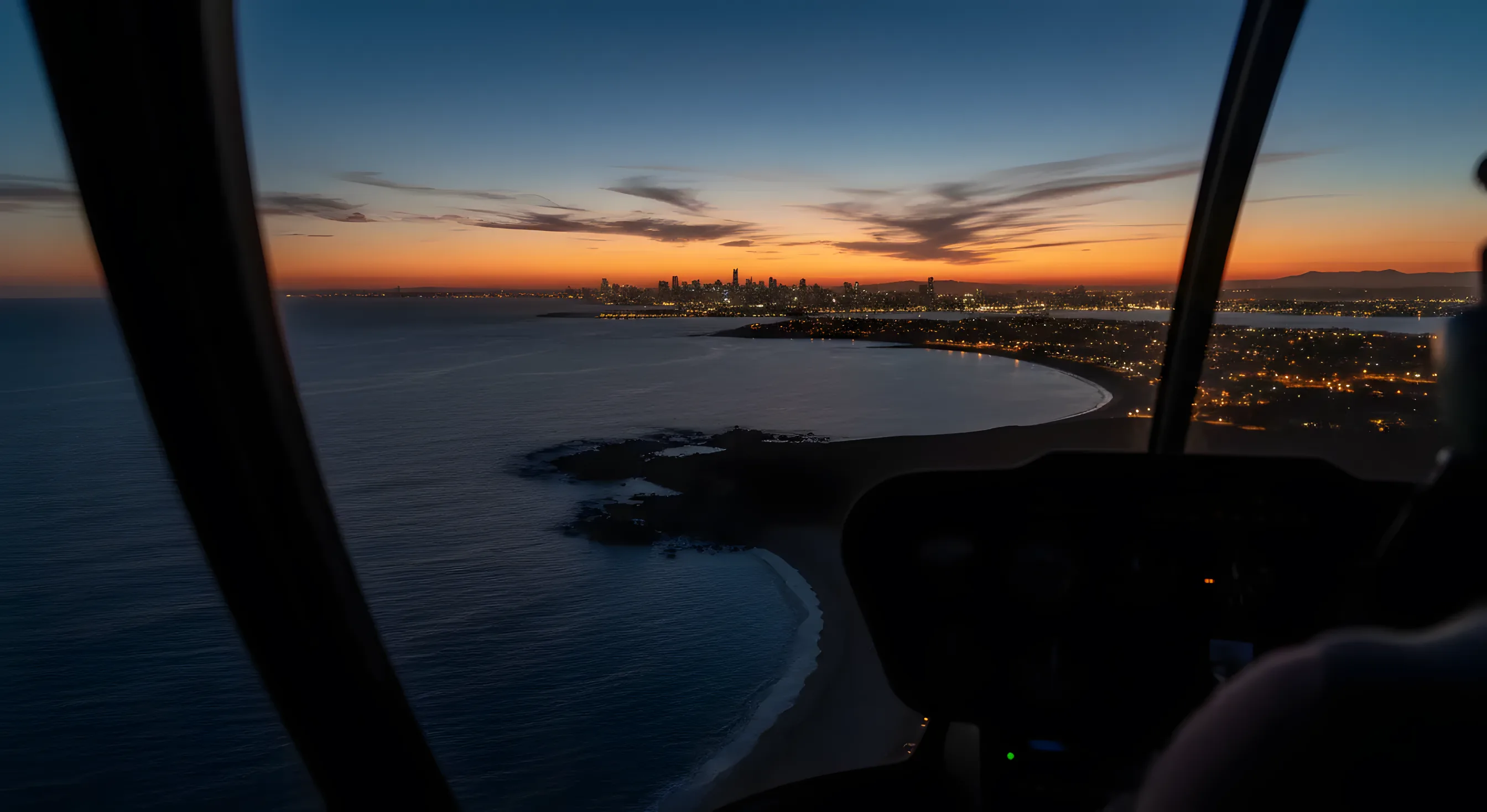 Aerial view of a coastline and a distant city skyline at dusk from inside a helicopter cockpit.