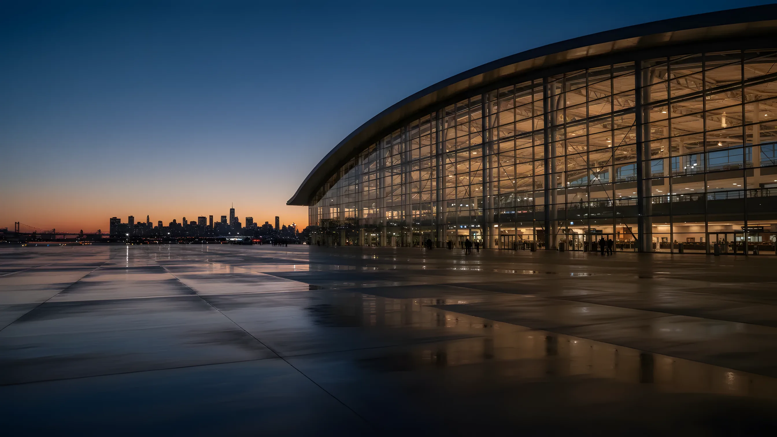 A modern airport terminal with a glass exterior glowing at dusk, with a city skyline in the background.
