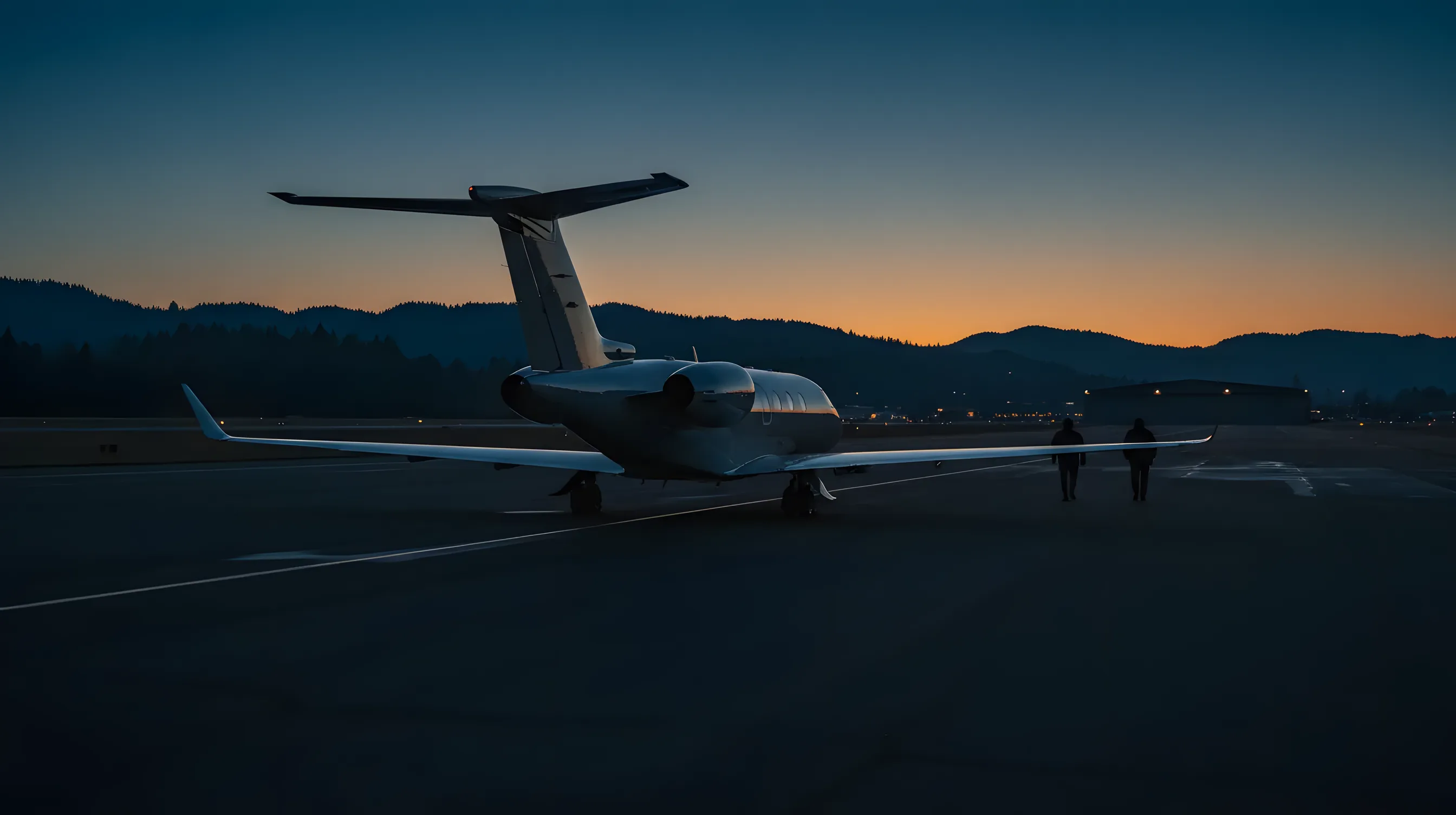 A private jet parked on an airfield at dusk, with forested hills and distant city lights in the background.