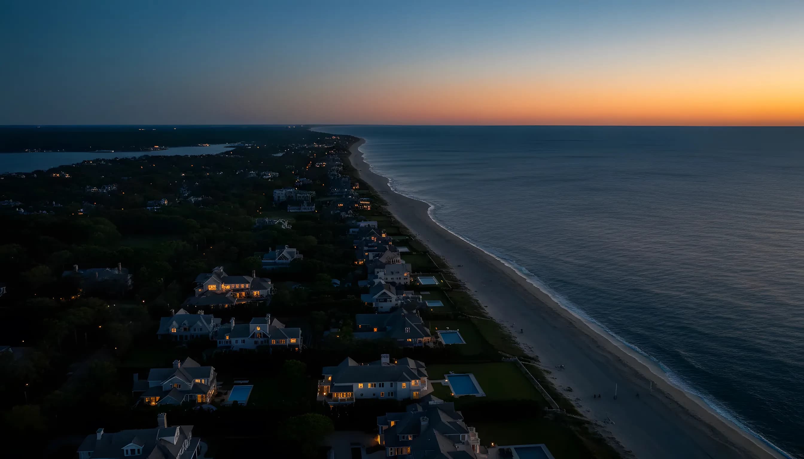 Aerial view of the Hamptons coastline at dusk, with lights from towns and homes glowing along the shore.