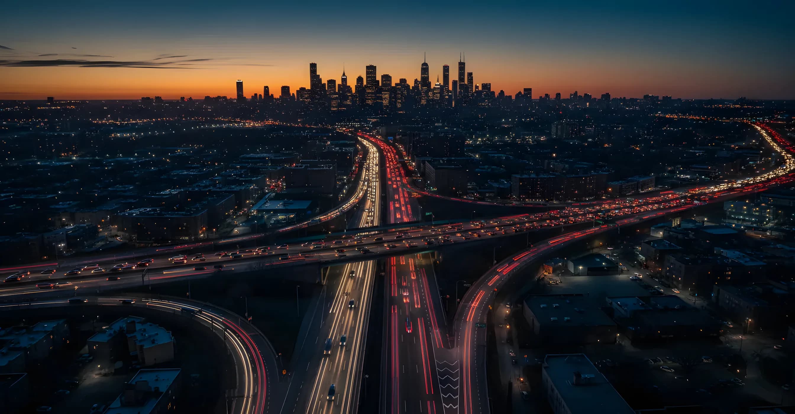 Aerial view of Queens, New York, at dusk with illuminated highways and the Manhattan skyline in the distance.