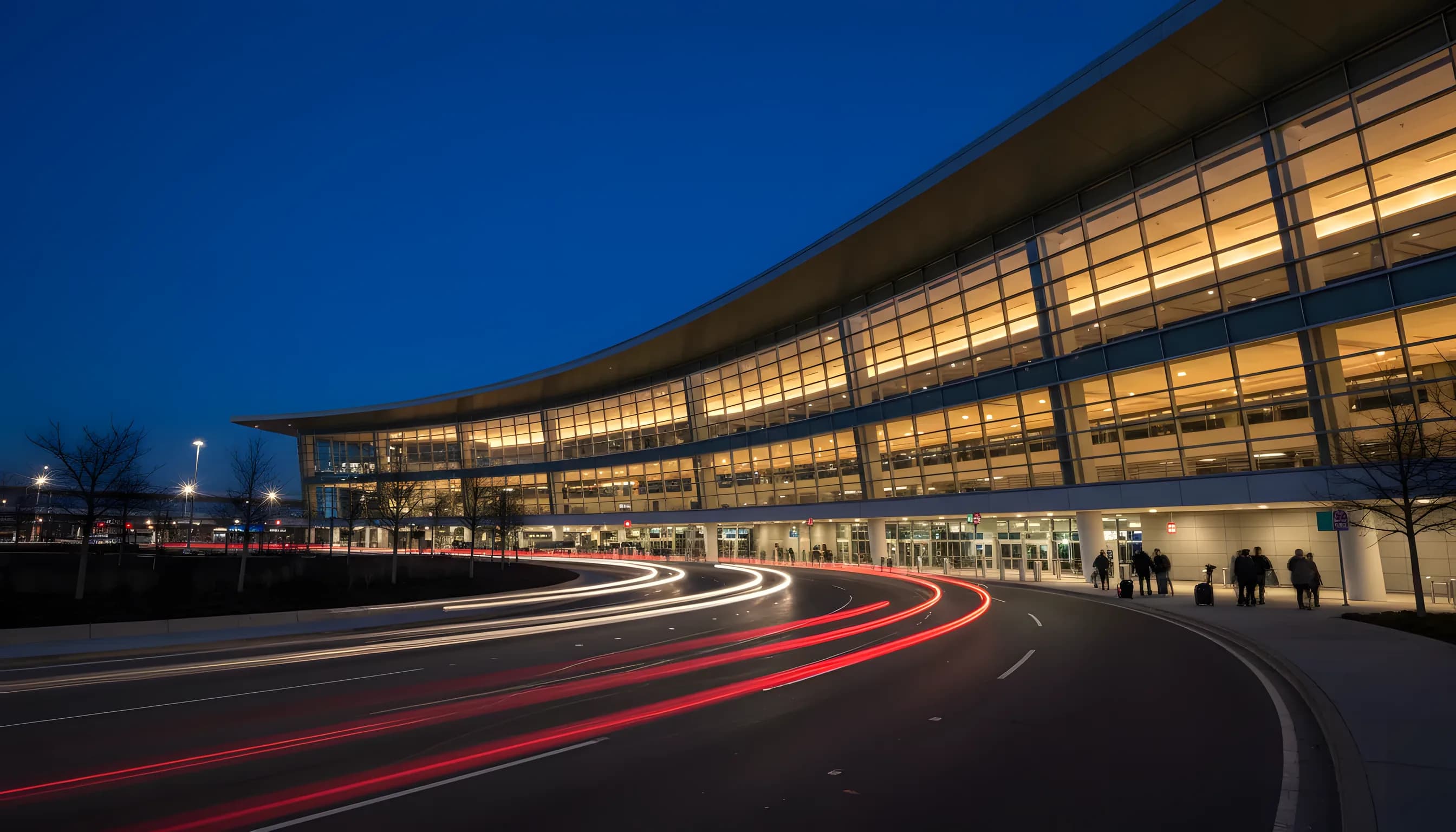 A modern glass airport terminal at dusk, with glowing lights and car light trails on the roadway in front.