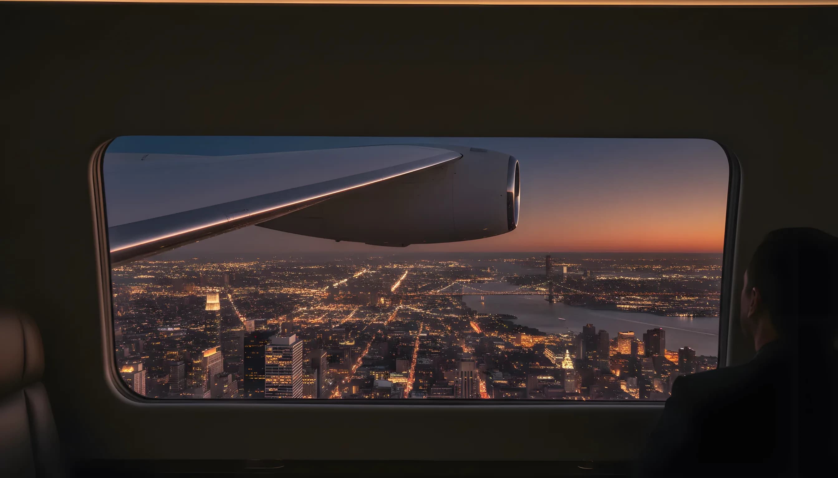 The wing of a private jet seen from a window, flying over the glowing lights of New York City at dusk.