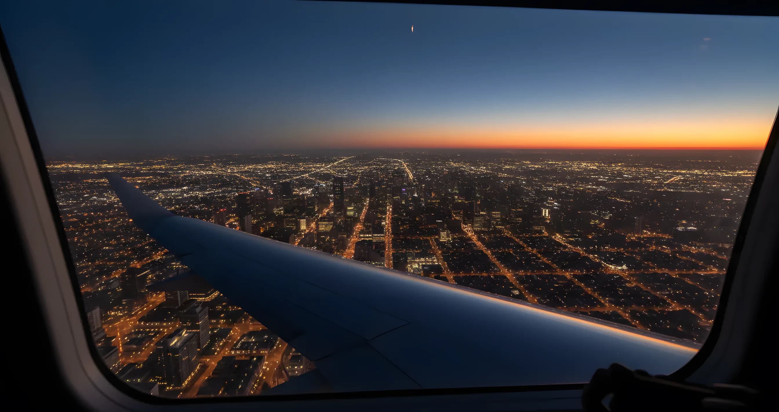 The wing of a private jet seen from the window, flying over a sprawling city with glowing lights at twilight.