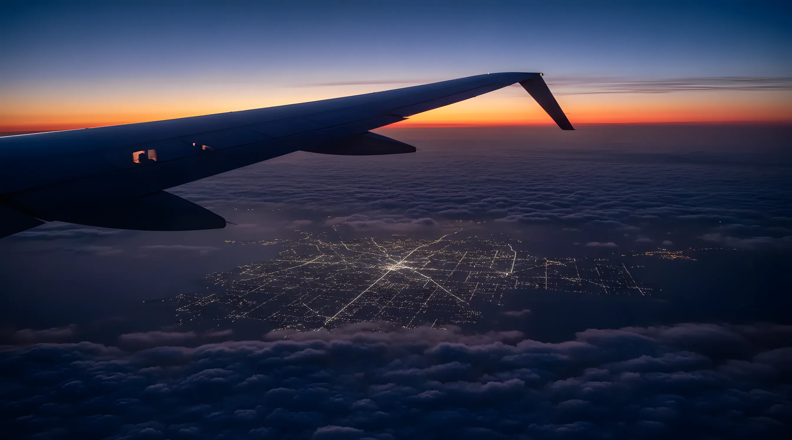 The wing of a jet flying high above clouds at dusk, with city lights visible far below.