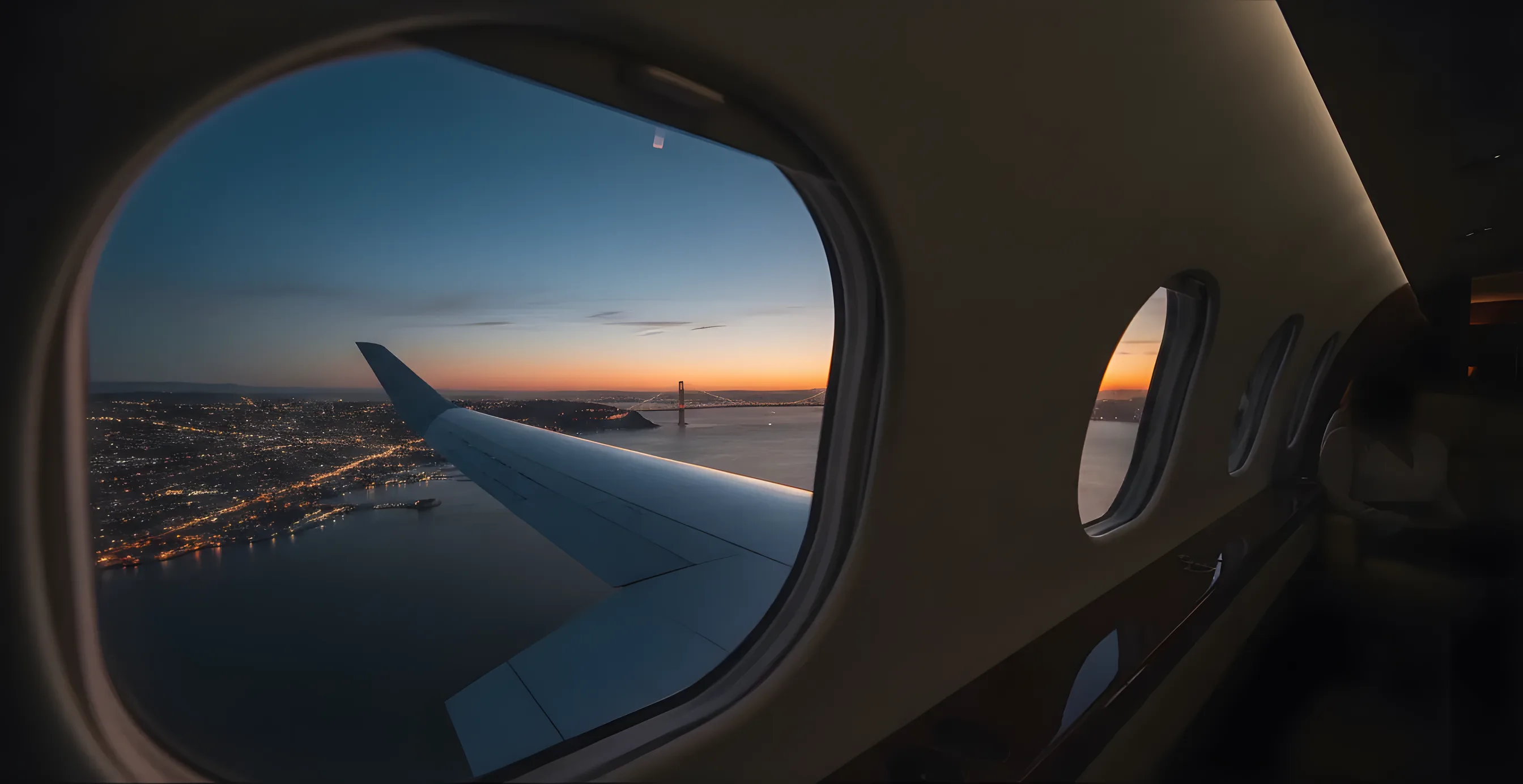 A private jet's wing seen from a window, flying over the San Francisco Bay at dusk with the Golden Gate Bridge in the distance.
