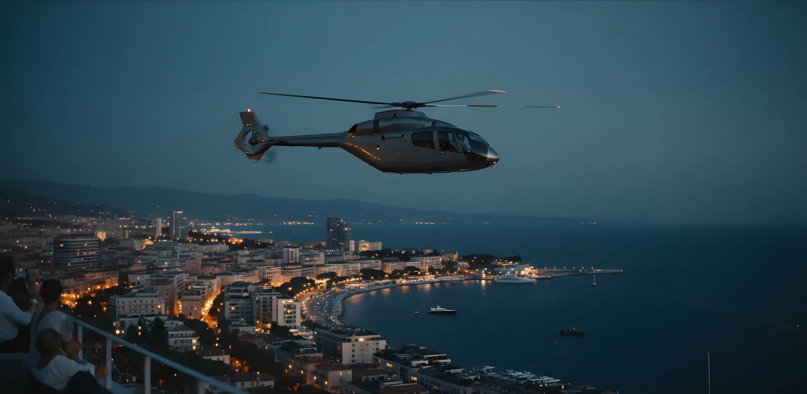 Un hélicoptère survole la Côte d'Azur au crépuscule, avec les lumières de Cannes en arrière-plan.