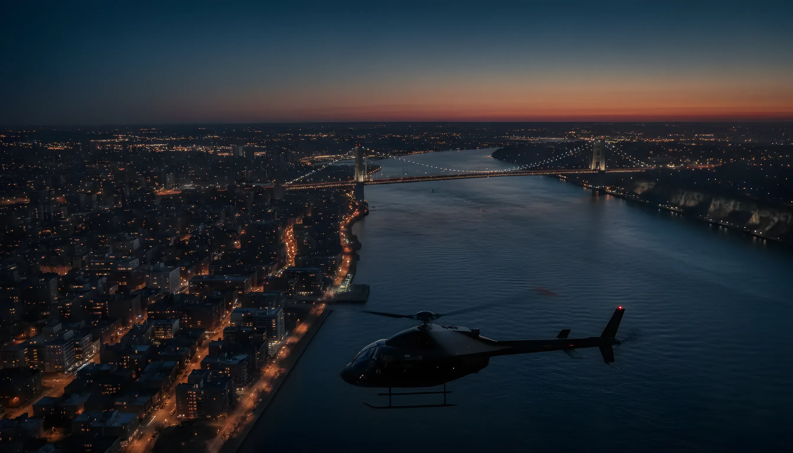 The George Washington Bridge lit up at dusk, with a helicopter flying over the Hudson River and Washington Heights in the background.