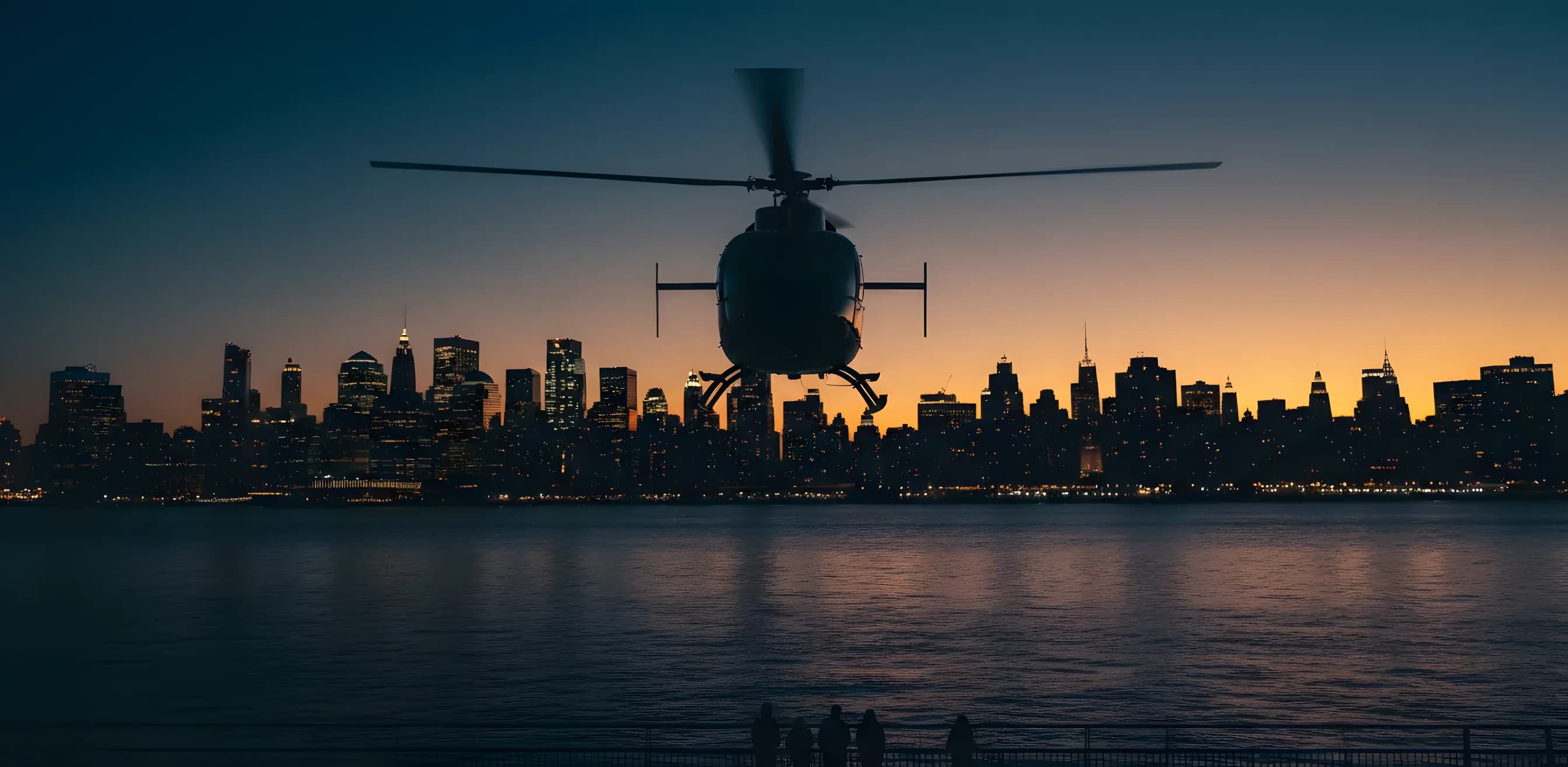 A helicopter flies over a river at dusk, with the illuminated New York City skyline in the background.