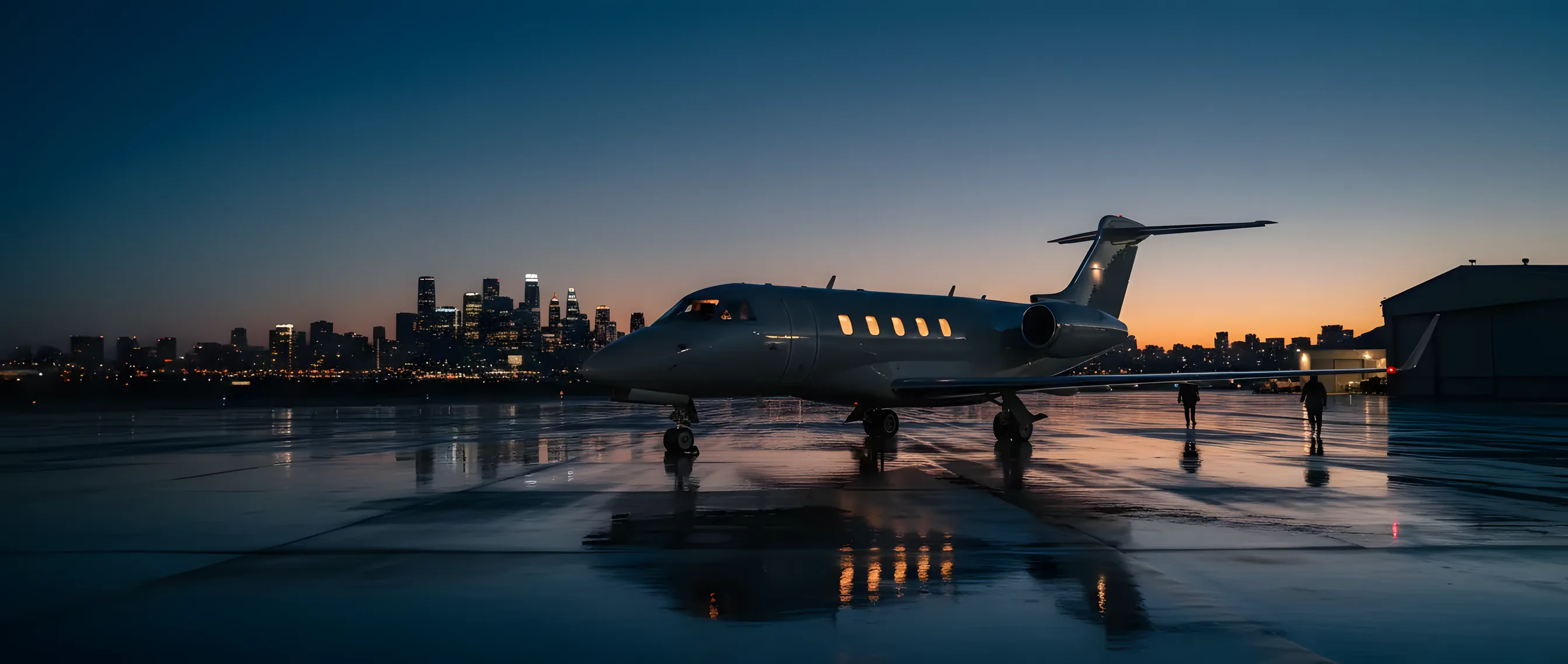 A private jet on an airport tarmac at dusk, with a city skyline in the background.