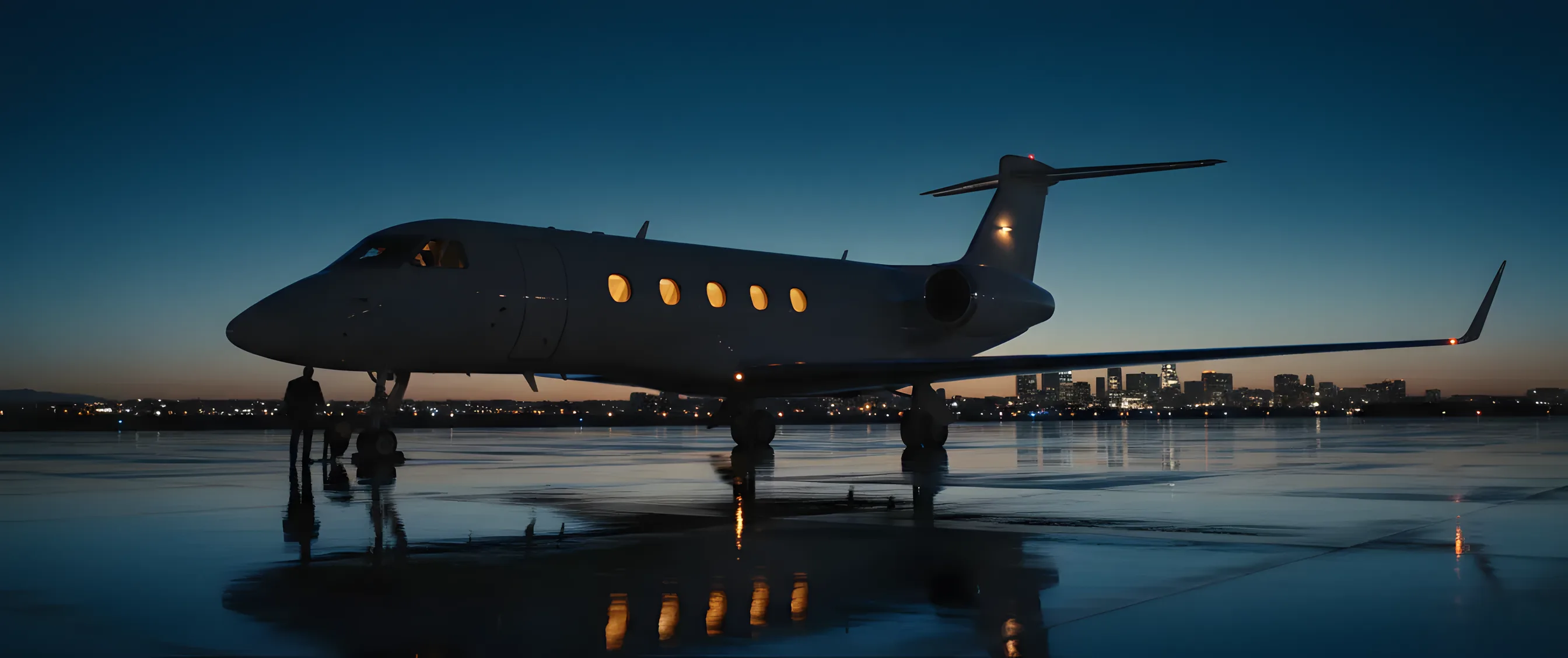 A private jet on an airfield at dusk, with its lights on and a city skyline in the background.