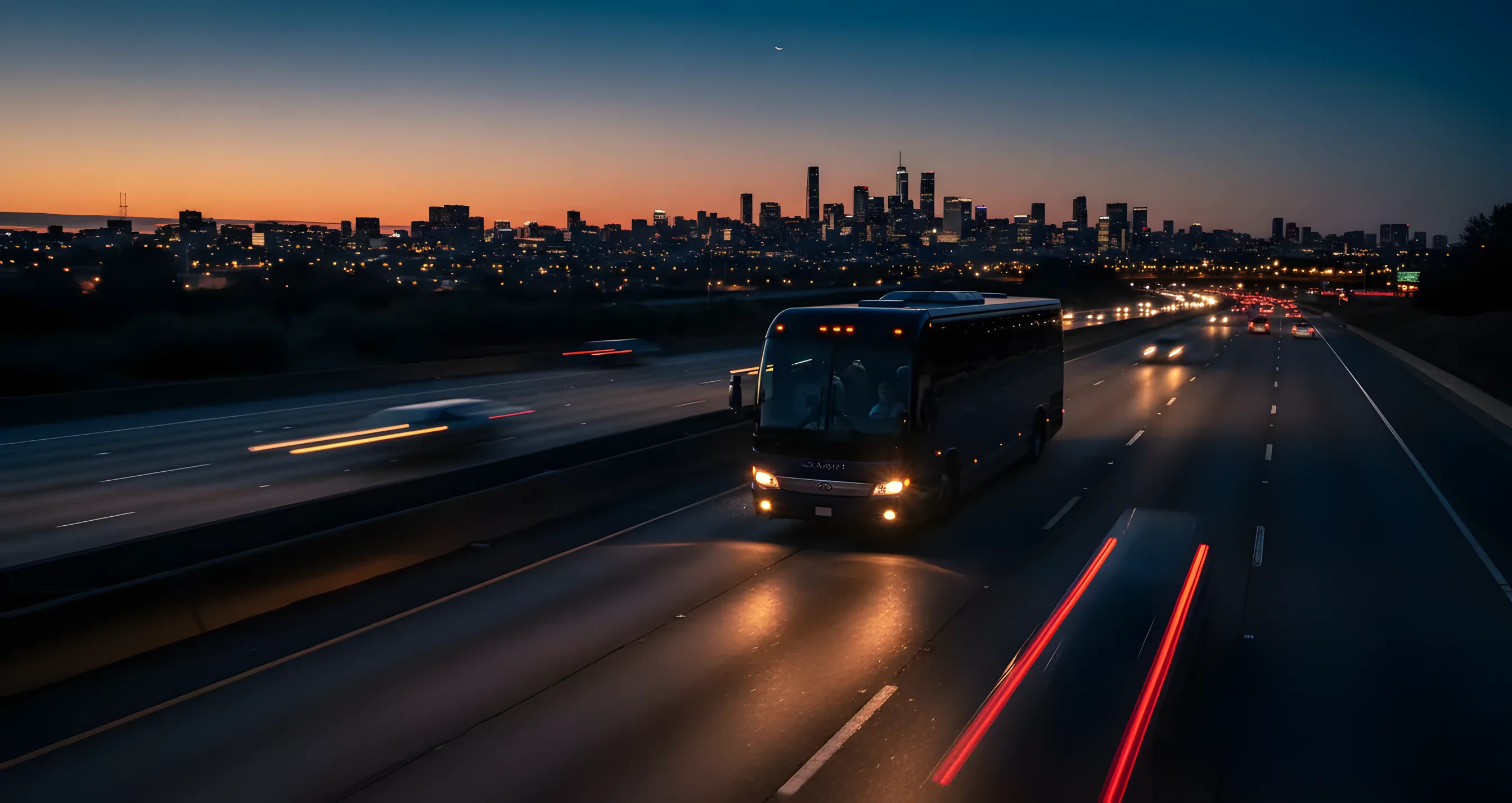 A luxury bus drives on a highway away from a city skyline at dusk.