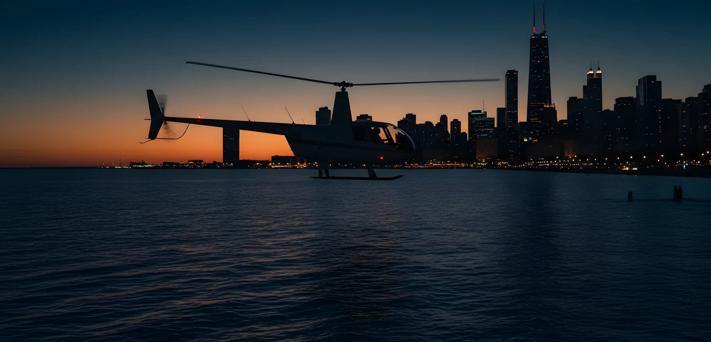 A helicopter flying over Lake Michigan with the Chicago skyline and Willis Tower behind it at blue hour.