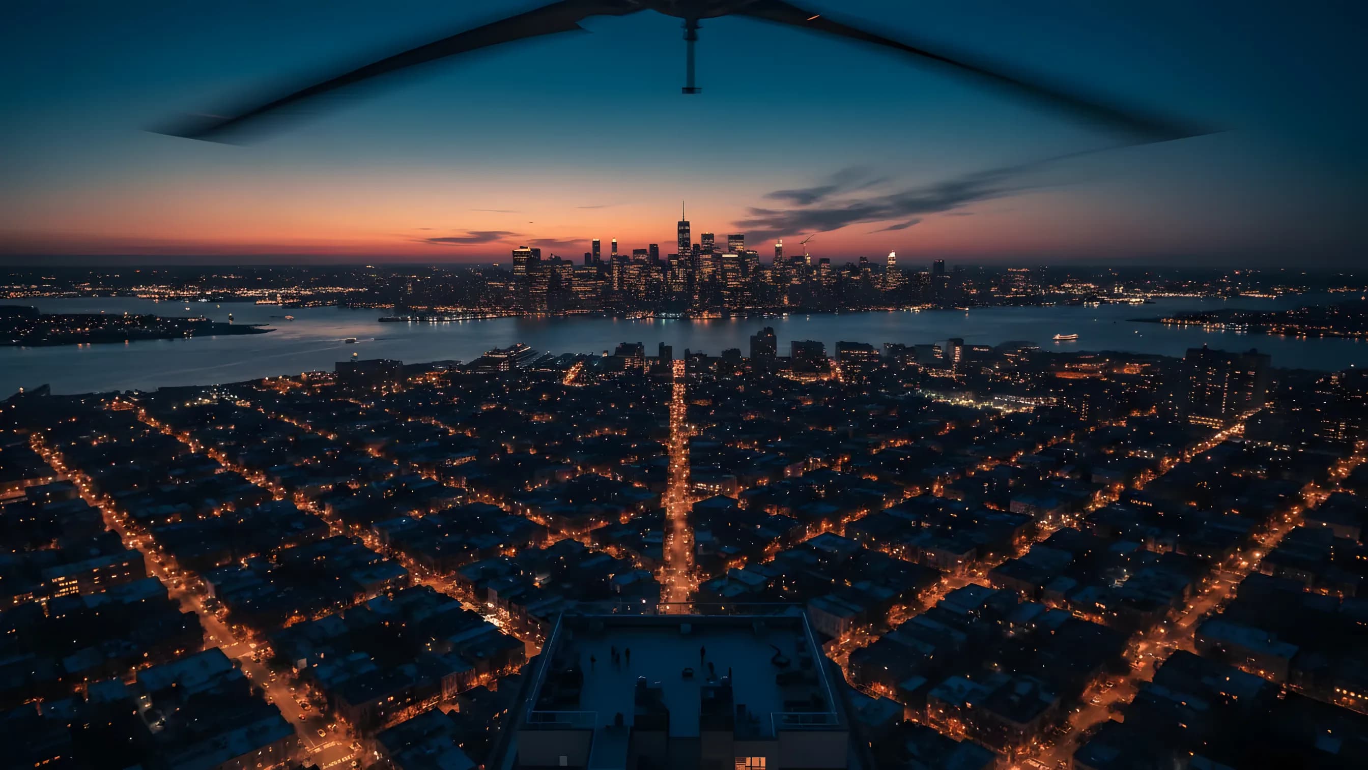 Aerial view at dusk of a historic neighborhood's glowing streets with the Manhattan skyline in the distance.