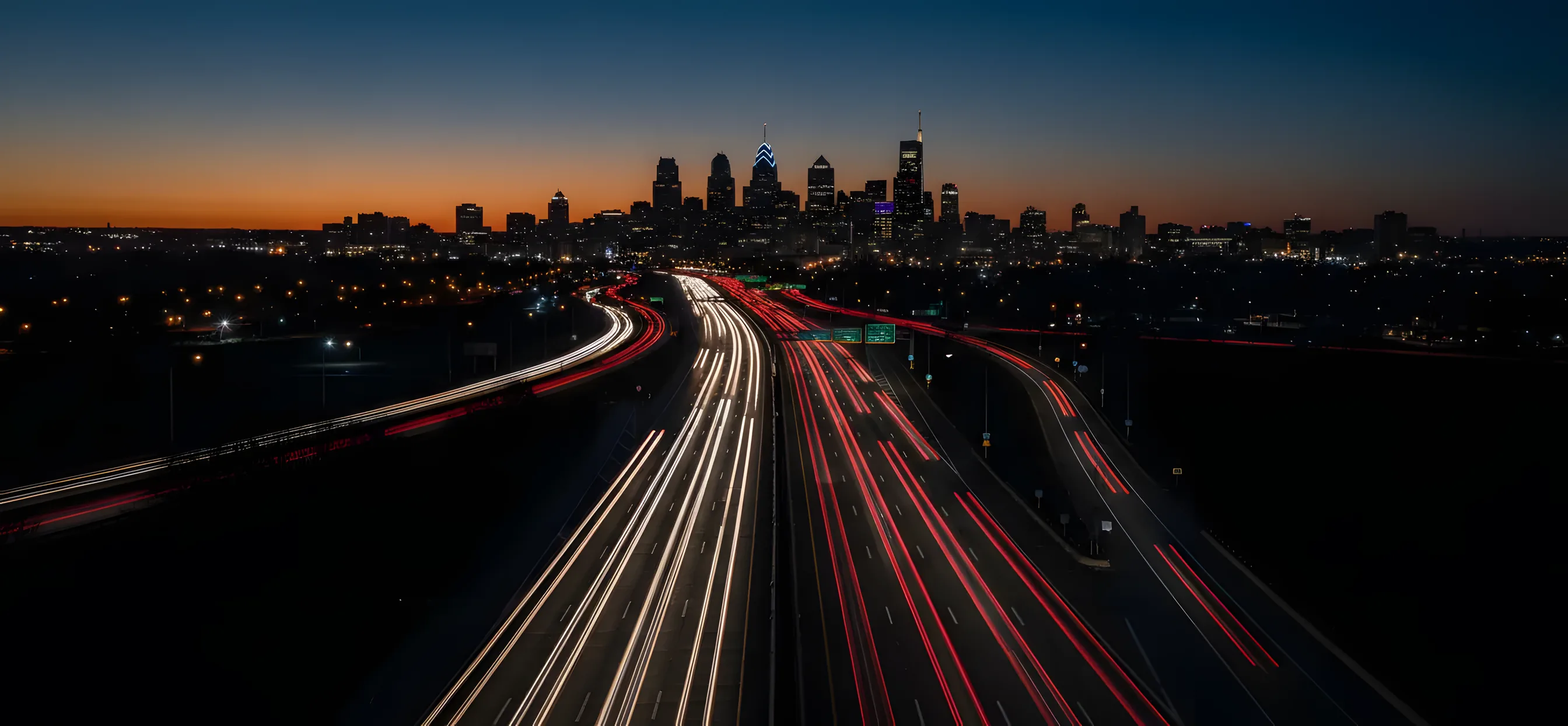 Aerial view of a highway at dusk with light trails from cars leading toward the Philadelphia skyline.