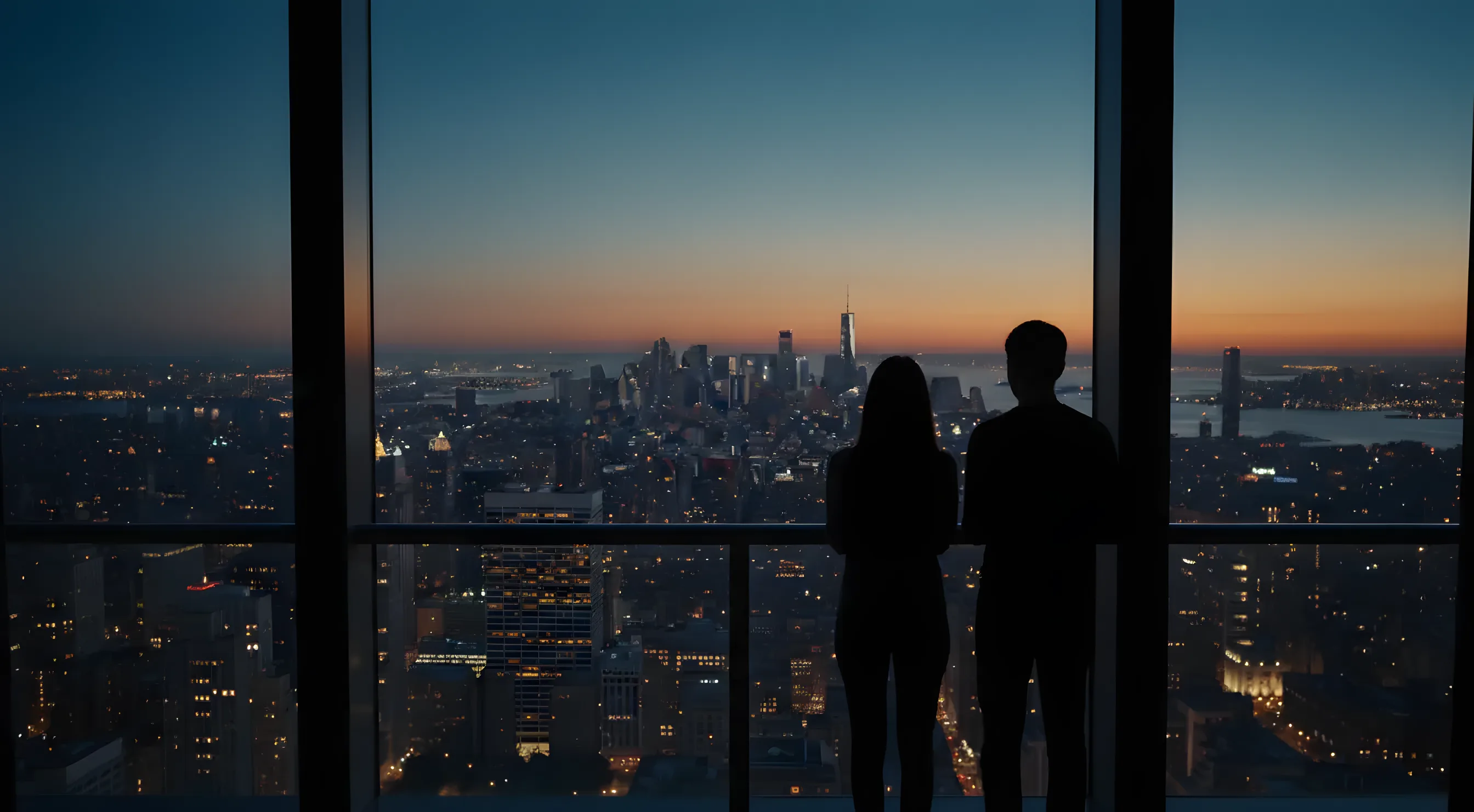 A couple looks out over the Manhattan skyline at dusk from a high-rise balcony.