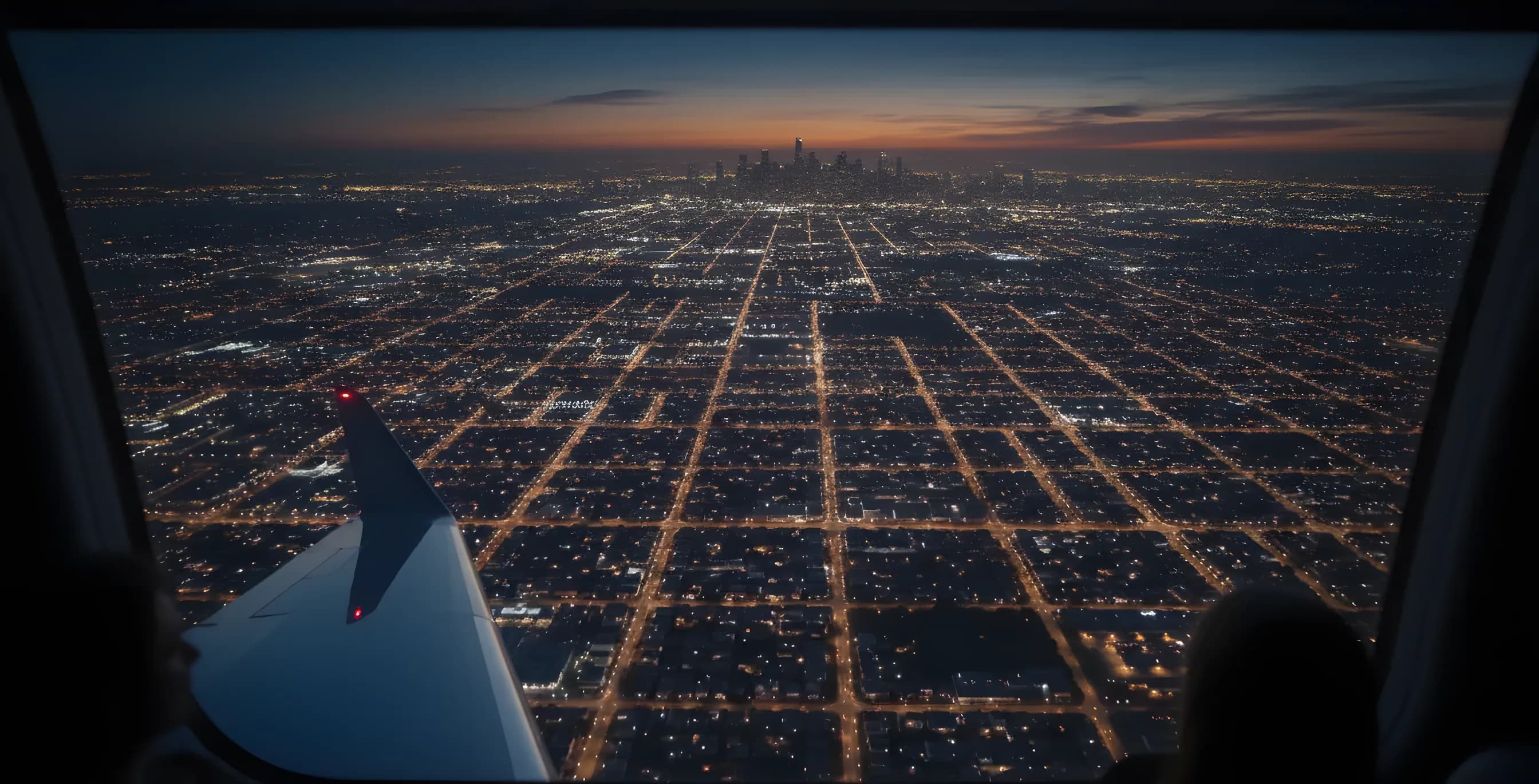 Aerial view from a plane window at dusk, showing glowing city lights below and an aircraft wingtip.