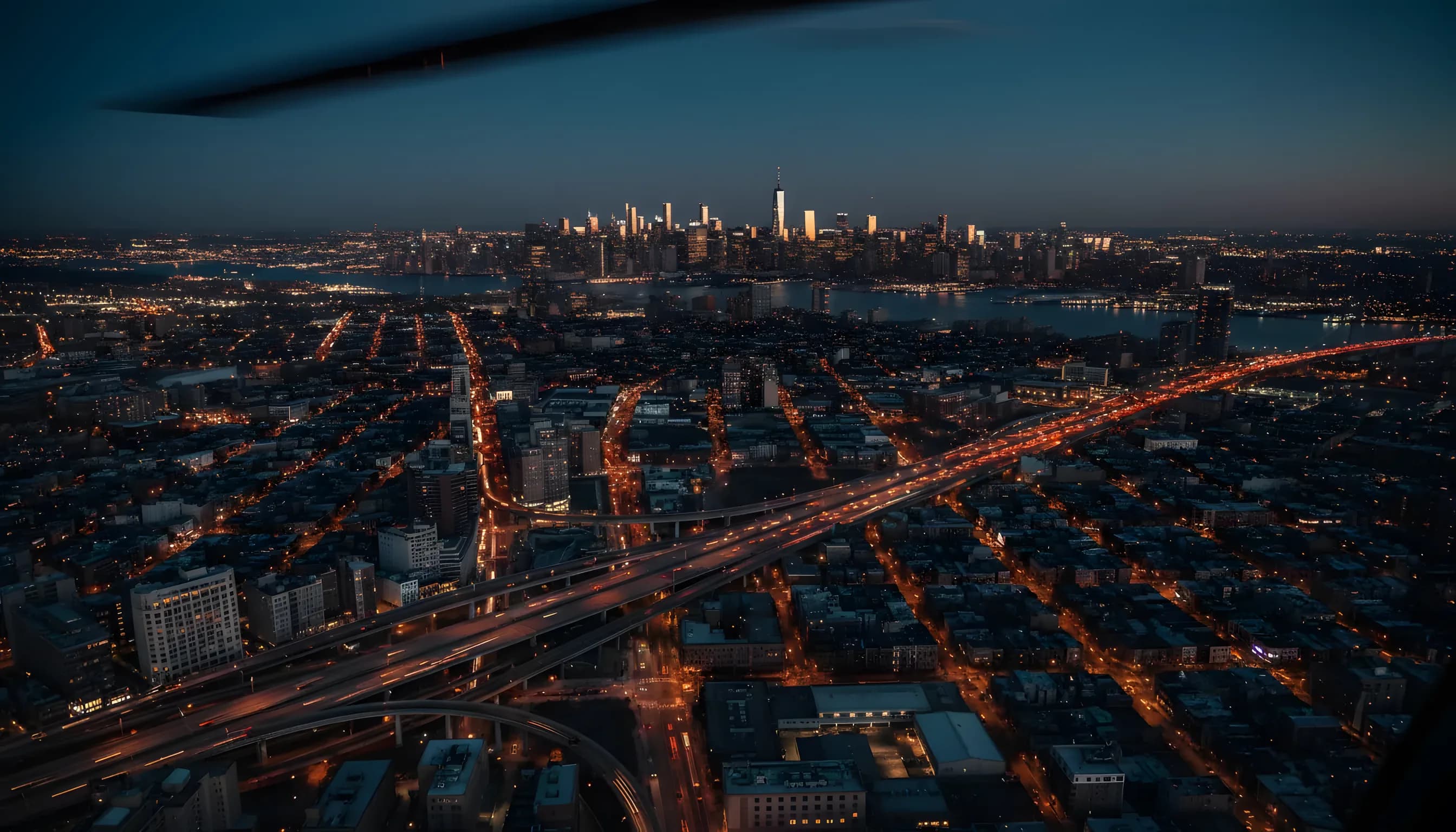 Aerial view of the Manhattan skyline at dusk, with city lights beginning to glow against a dark blue sky.