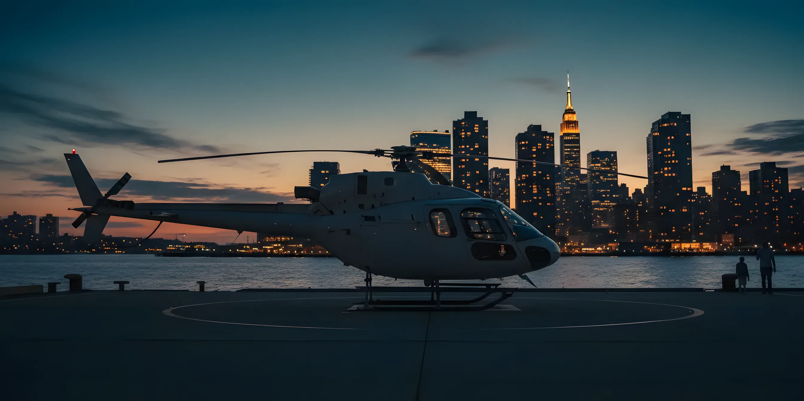 A helicopter on a Hudson River heliport pier at blue hour with Hudson Yards and the Empire State Building behind.
