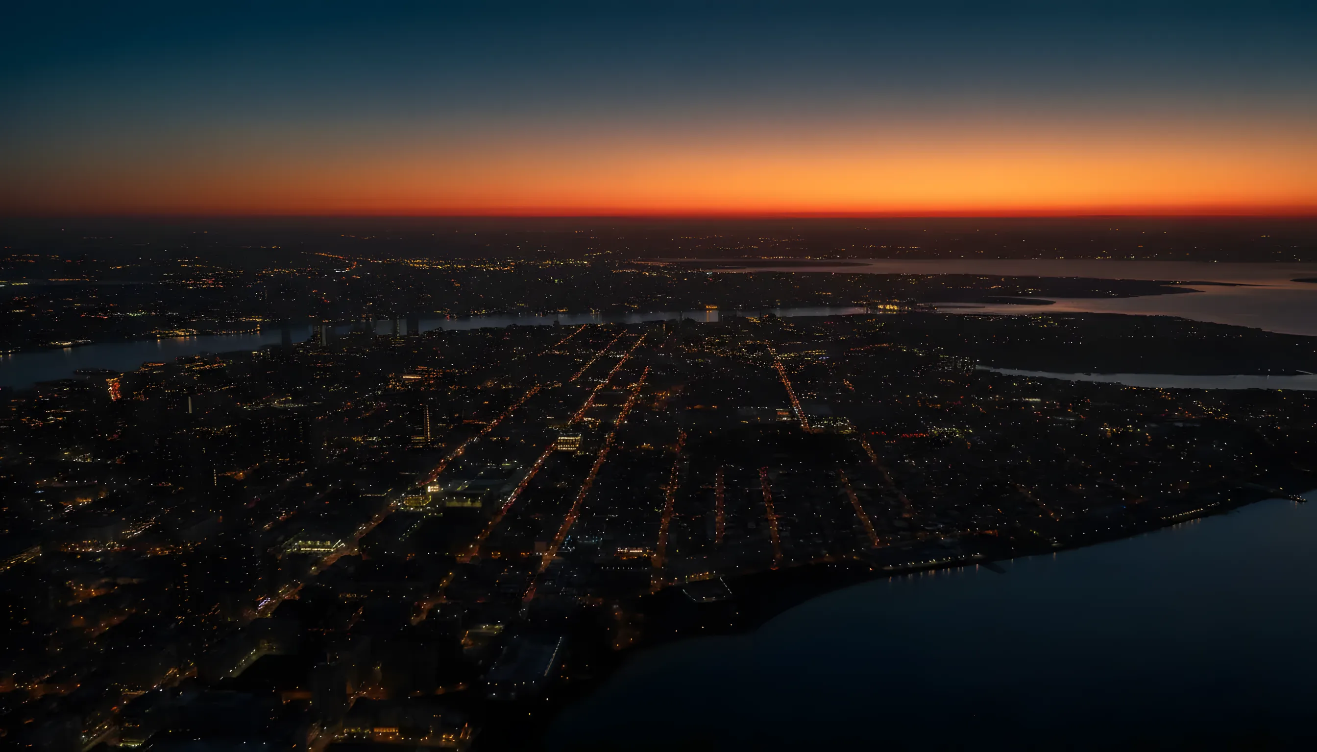Aerial view from a helicopter at dusk, flying away from a glowing city skyline towards the coast.