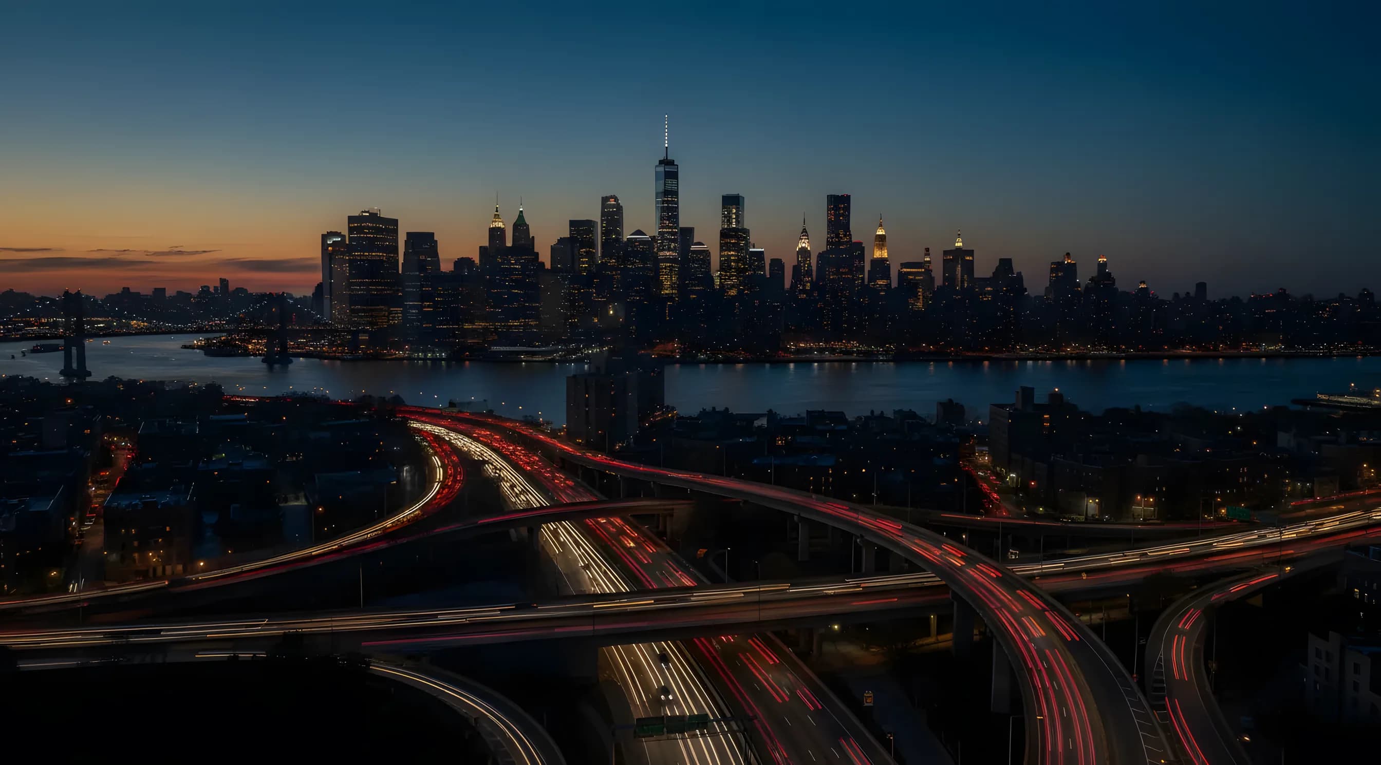 Aerial view of Queens, New York at dusk, with traffic on the highways and the Manhattan skyline in the distance.