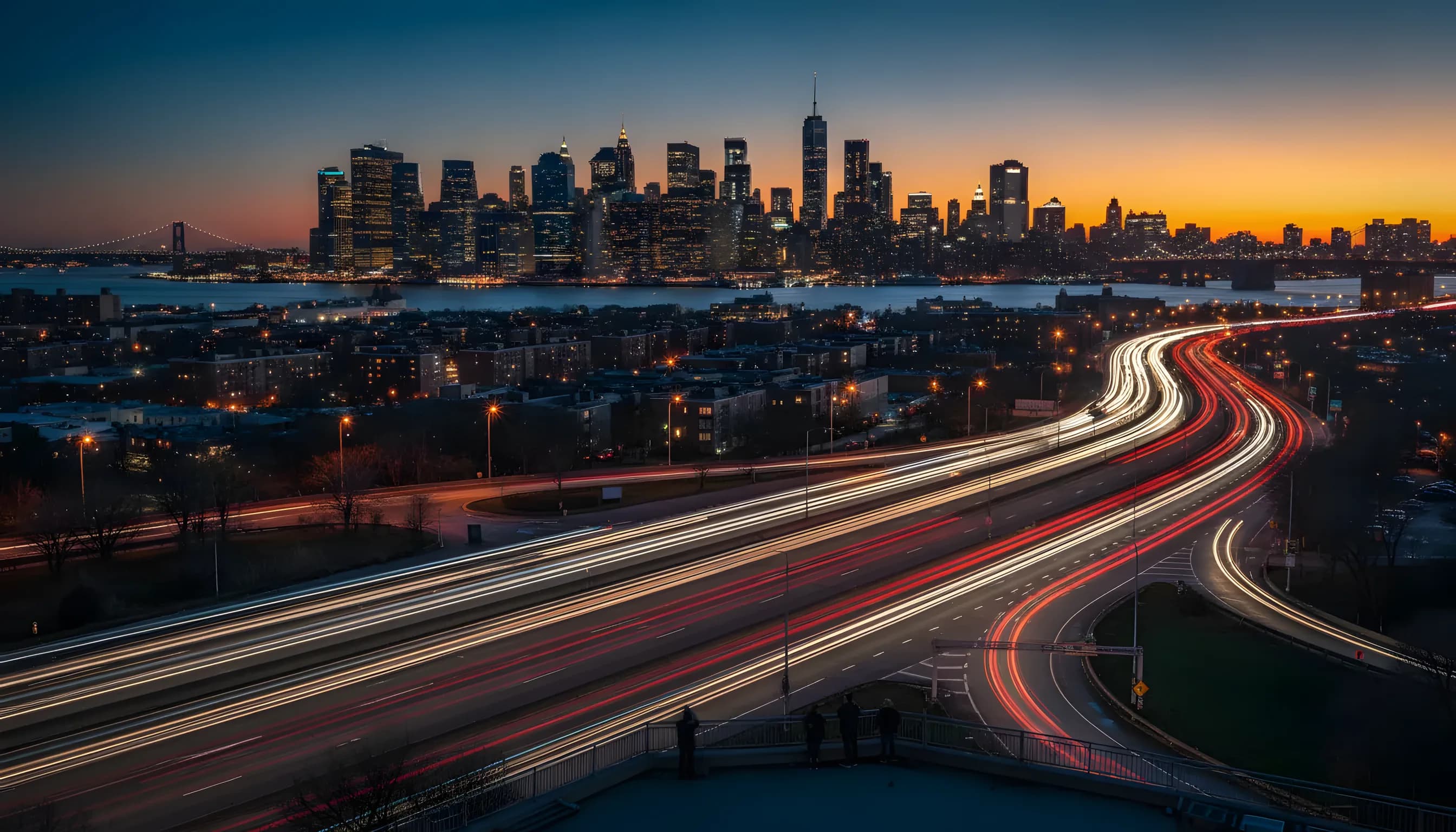The Manhattan skyline seen from Queens at dusk, with light trails from traffic on a highway in the foreground.