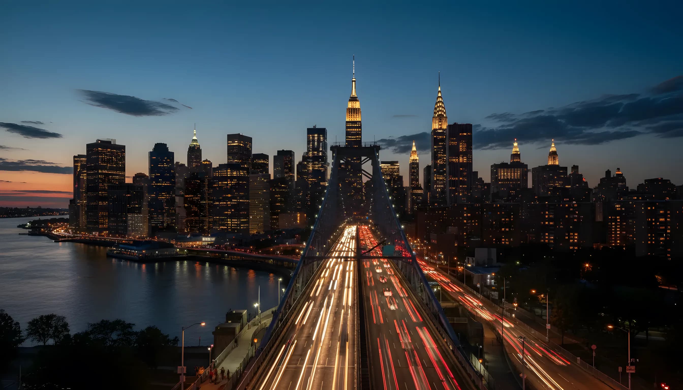The Manhattan skyline glows at dusk, with streaks of light from traffic on a bridge crossing the East River.
