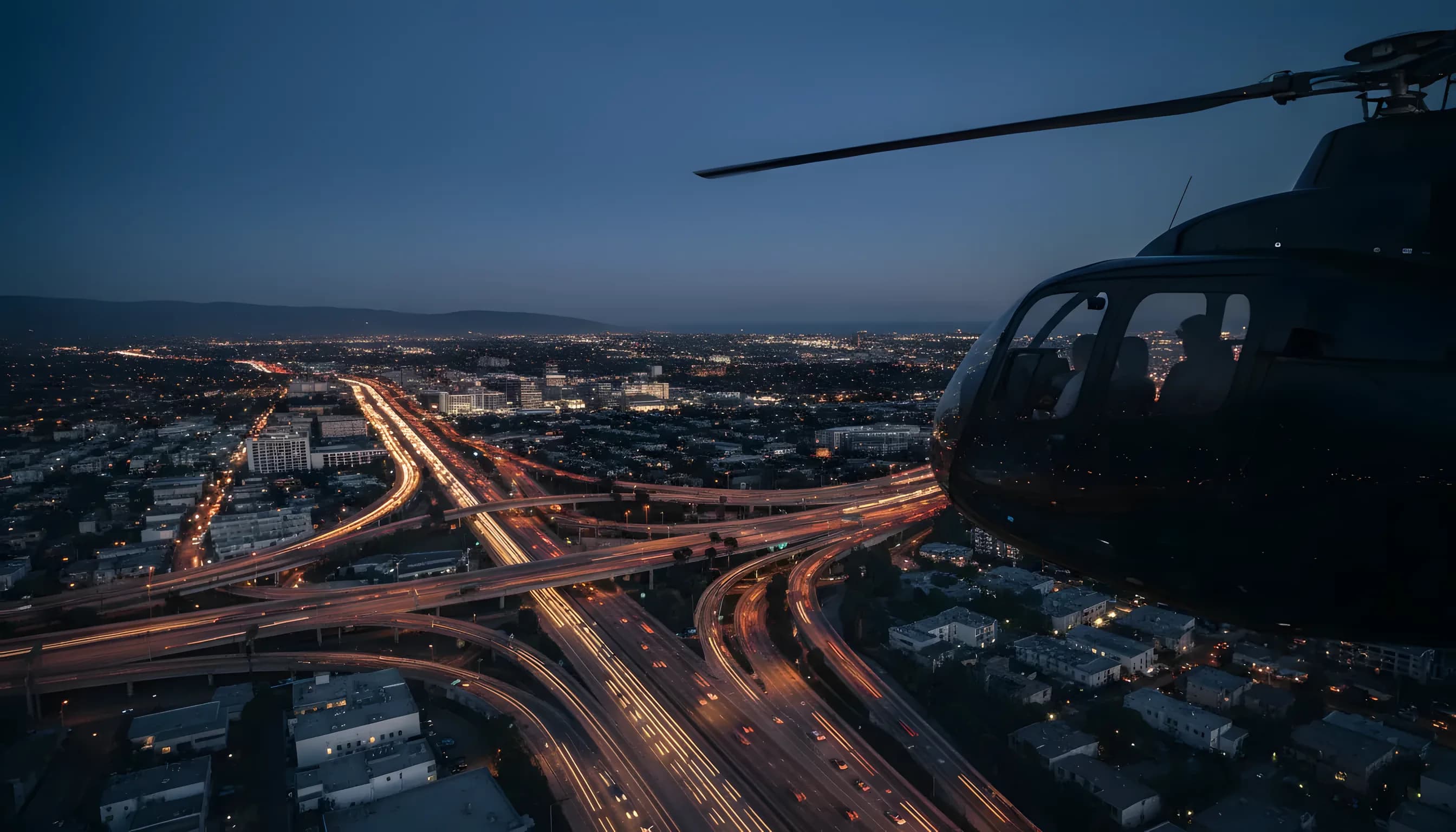 A helicopter soars over the vast, illuminated Los Angeles cityscape at dusk.