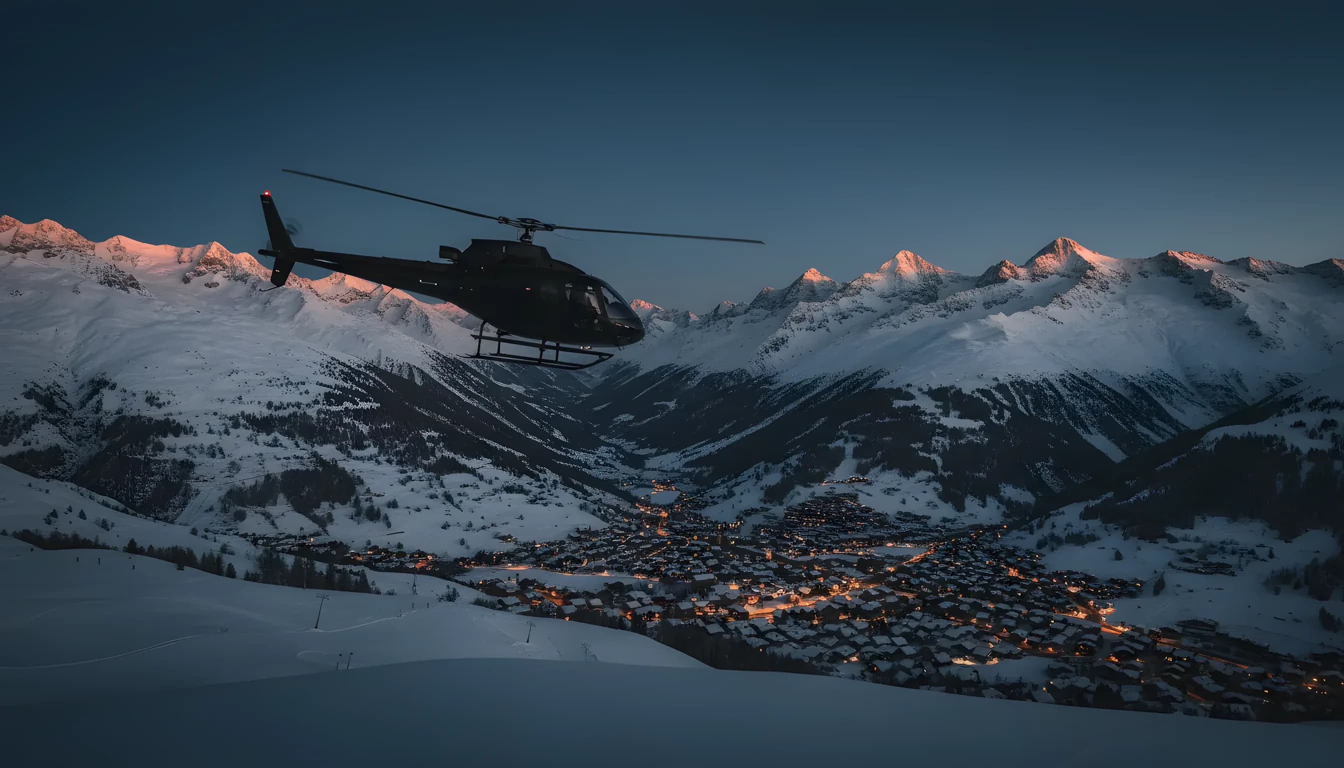 A helicopter flies over a snowy mountain valley toward a glowing alpine village at dusk.