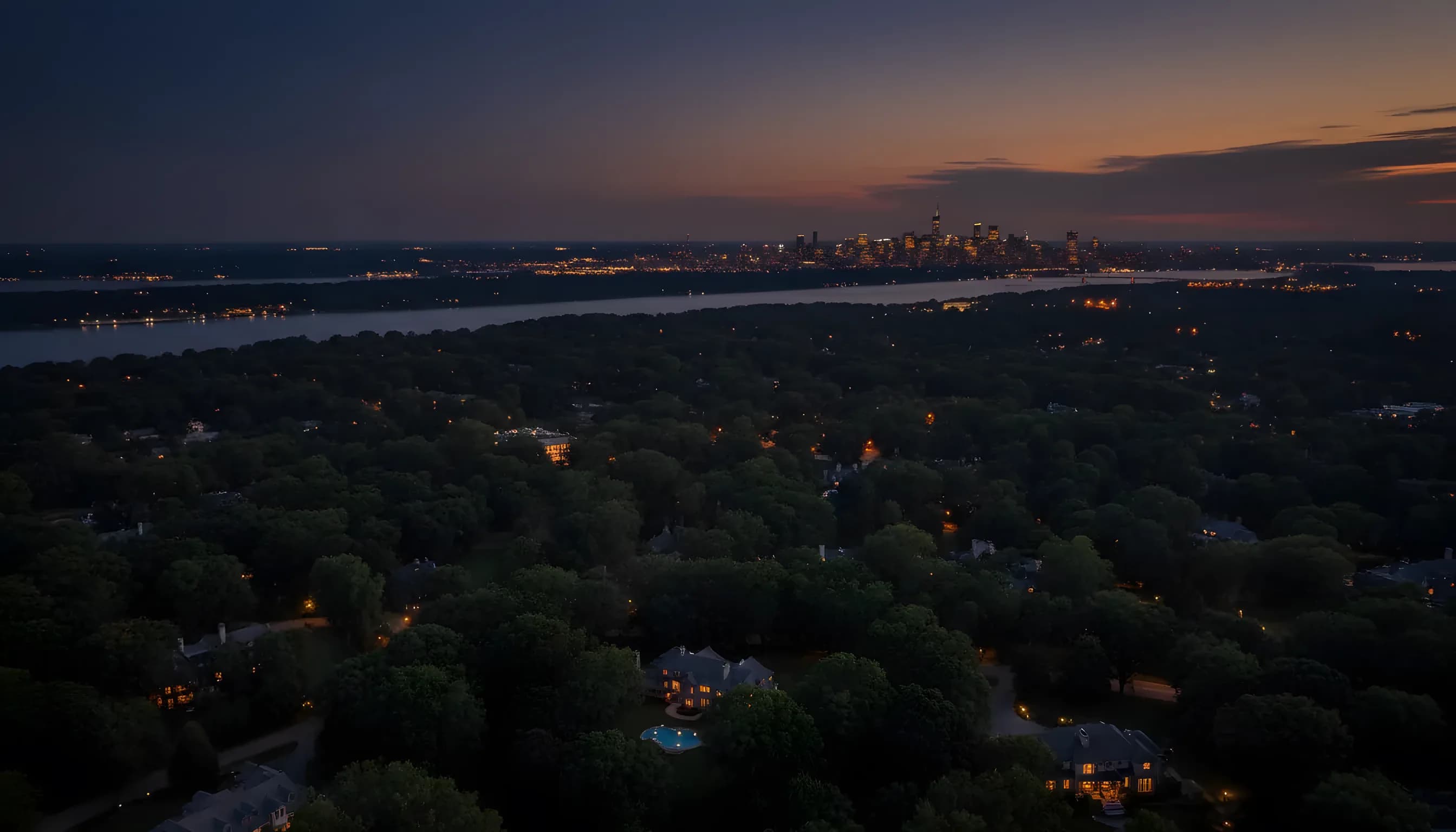 A view over the Hudson River from Westchester at dusk, with the distant lights of the New York City skyline on the horizon.