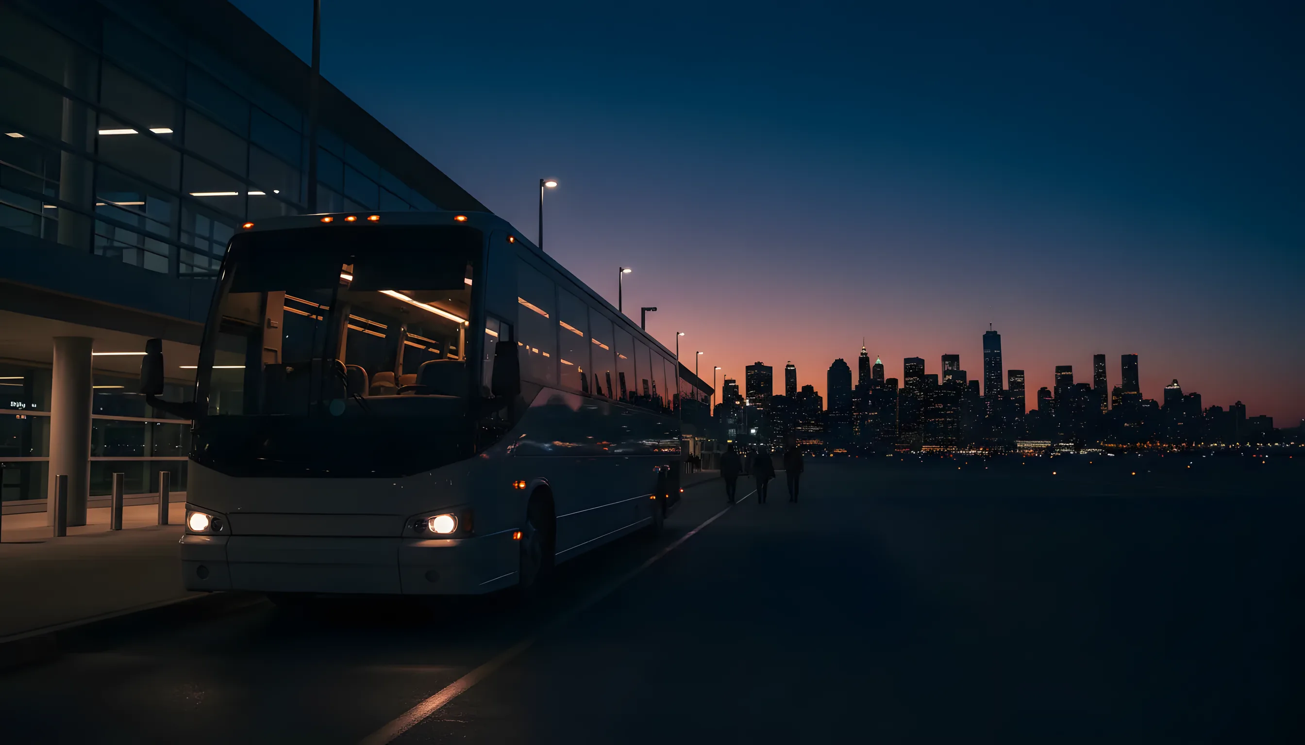 A modern charter bus at an airport terminal curb at dusk, with a city skyline glowing in the distance.