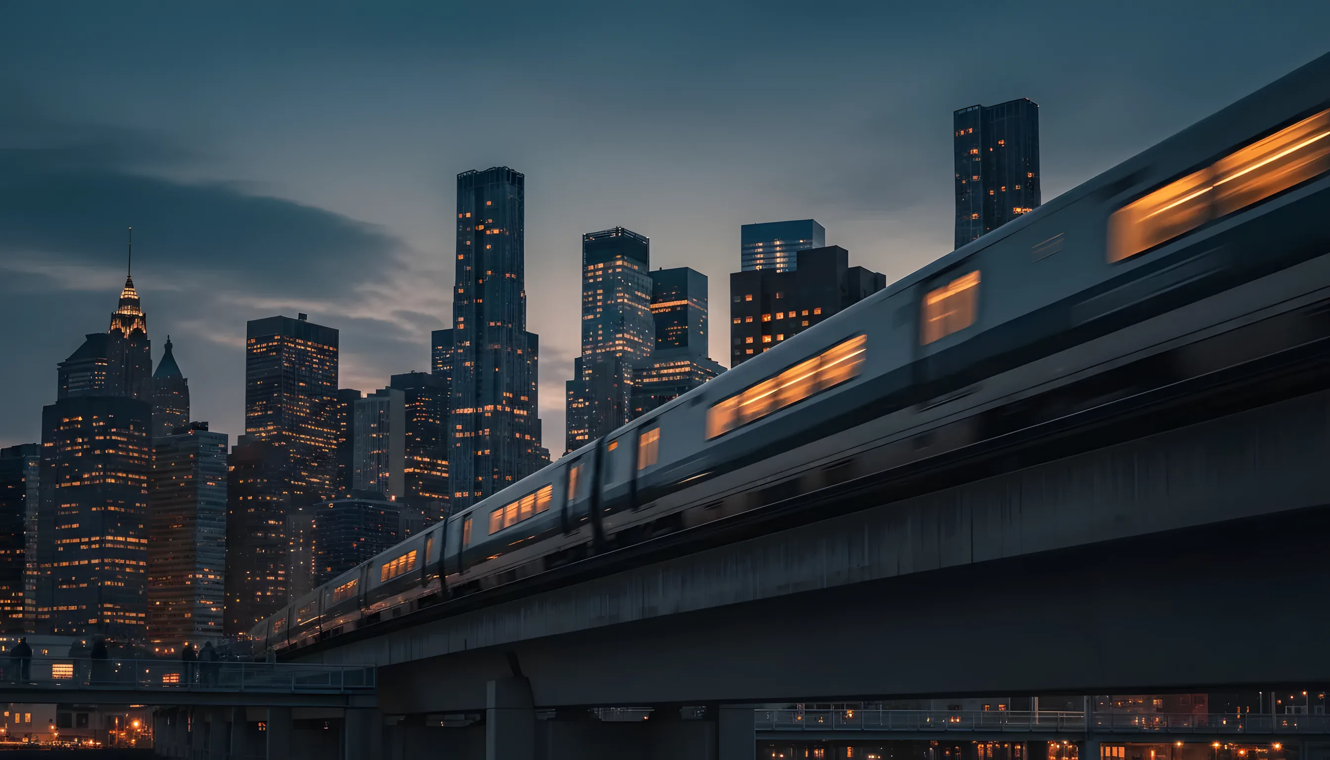 A modern train speeds past at dusk, with the illuminated Lower Manhattan skyline visible in the distance.