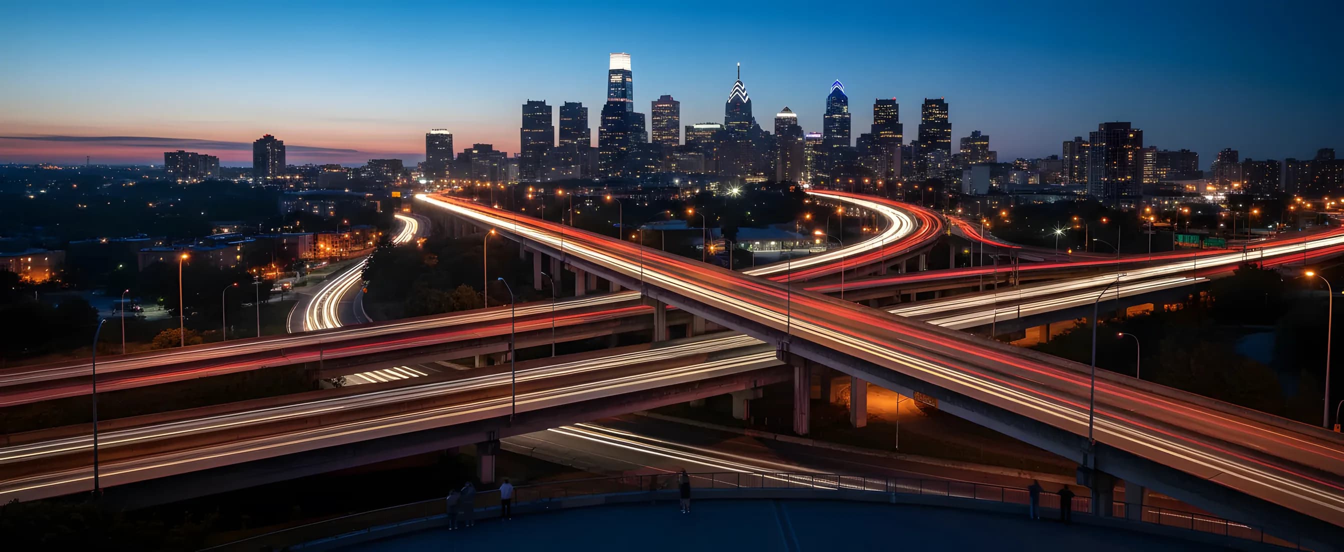 Light trails from traffic on a highway leading toward the Philadelphia skyline at dusk.
