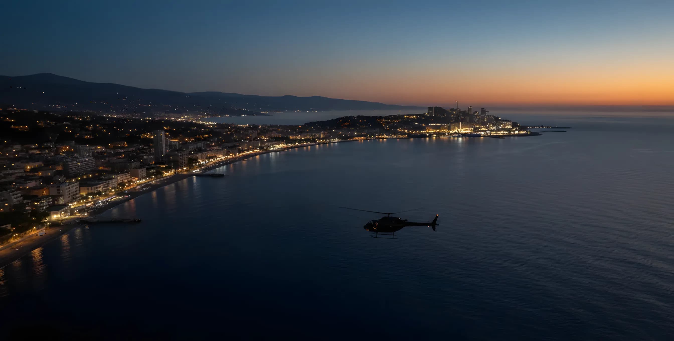 An aerial view of the French Riviera coastline at dusk, with a helicopter flying towards the lights of Monaco.