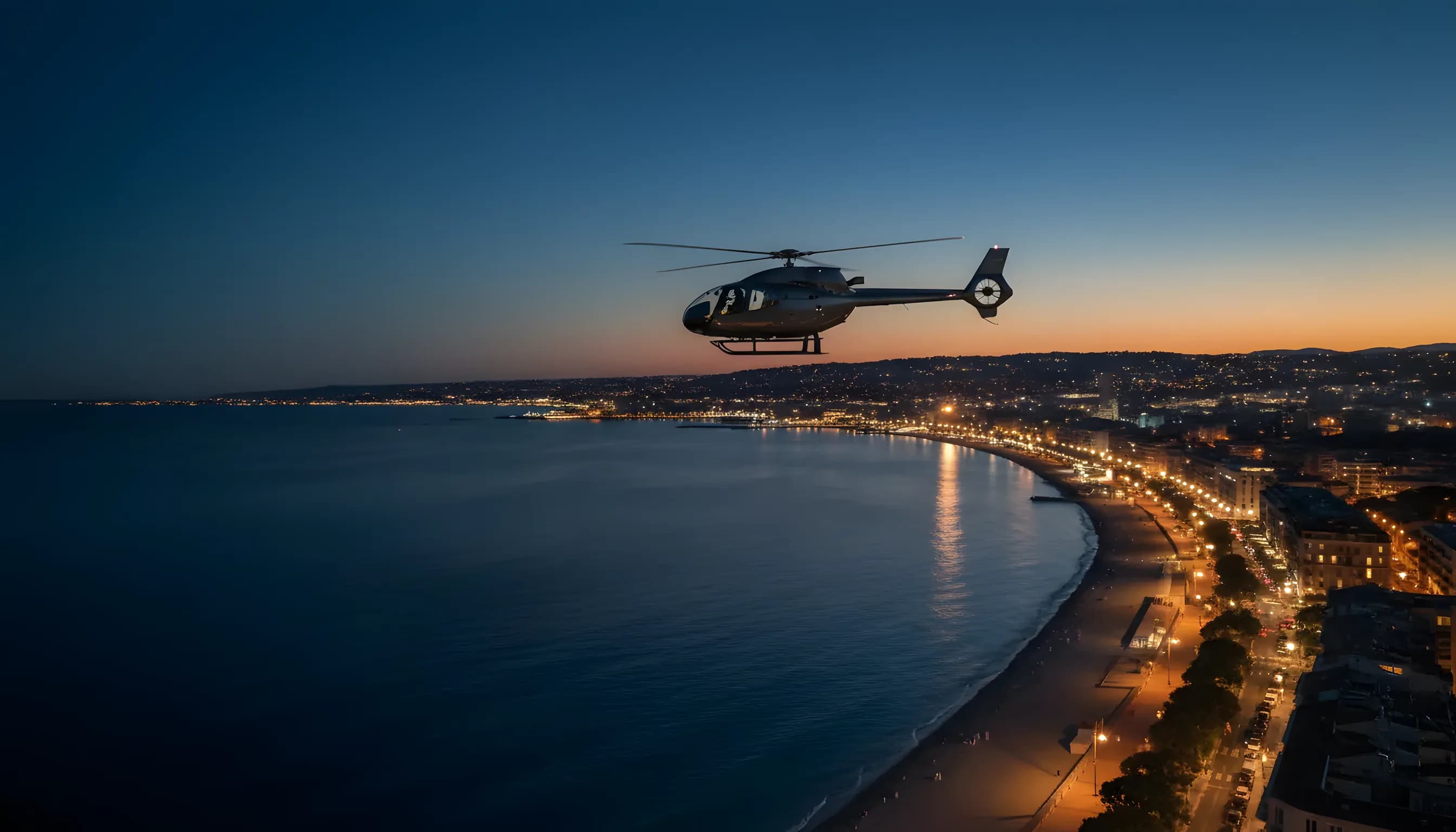 A helicopter flies along the French Riviera coastline at twilight, with the lights of Cannes glowing in the distance.