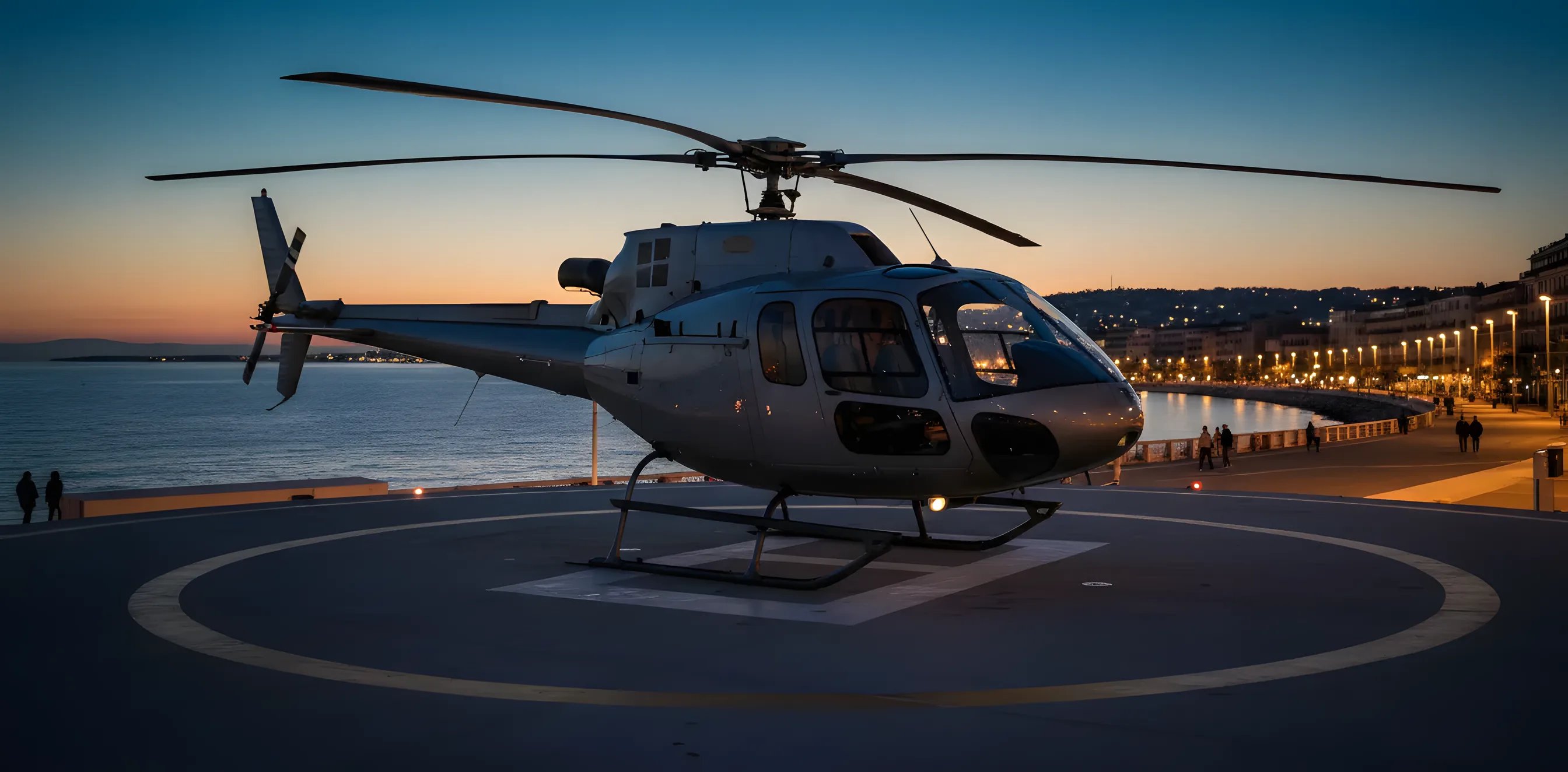 A helicopter on a seaside helipad in Nice at twilight with the Promenade des Anglais behind it.