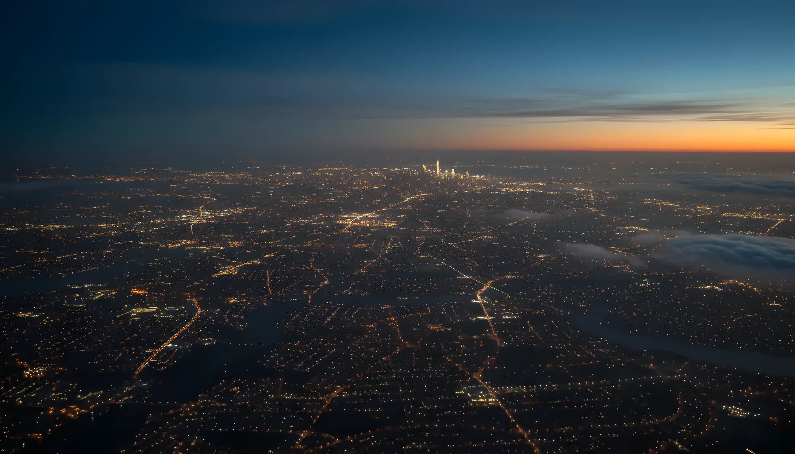Aerial view from an aircraft at dusk, with the lights of a city and landscape stretching out below.