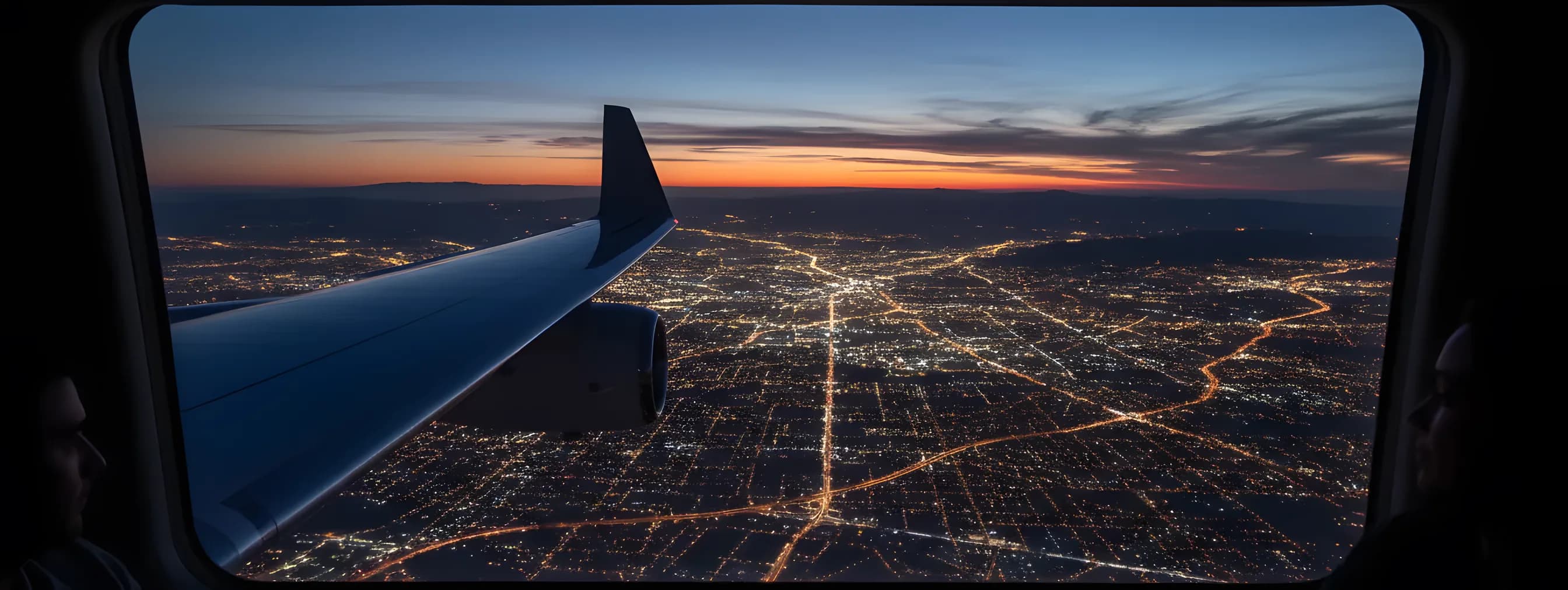 View from a private jet window at dusk, showing the wing over a glowing city grid below.