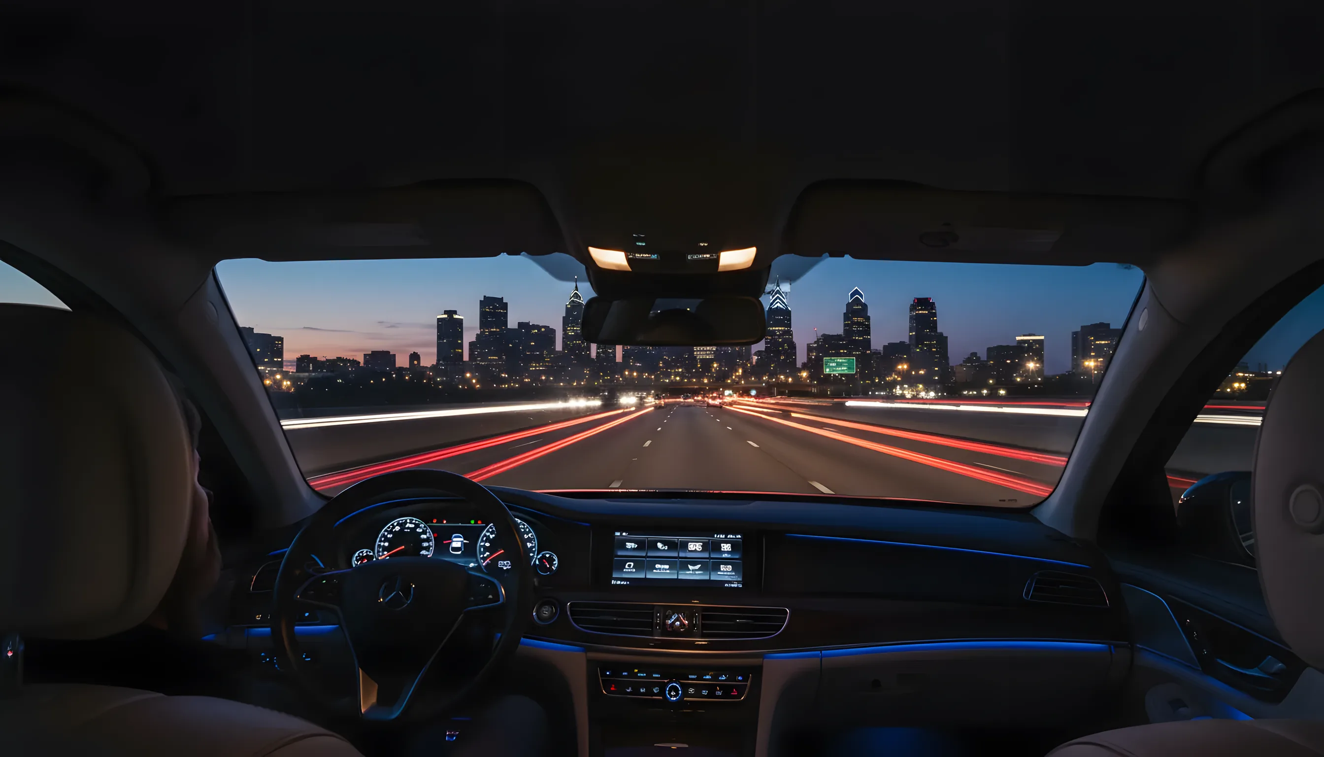 The Philadelphia skyline at dusk with glowing city lights, viewed from a moving car on a highway.